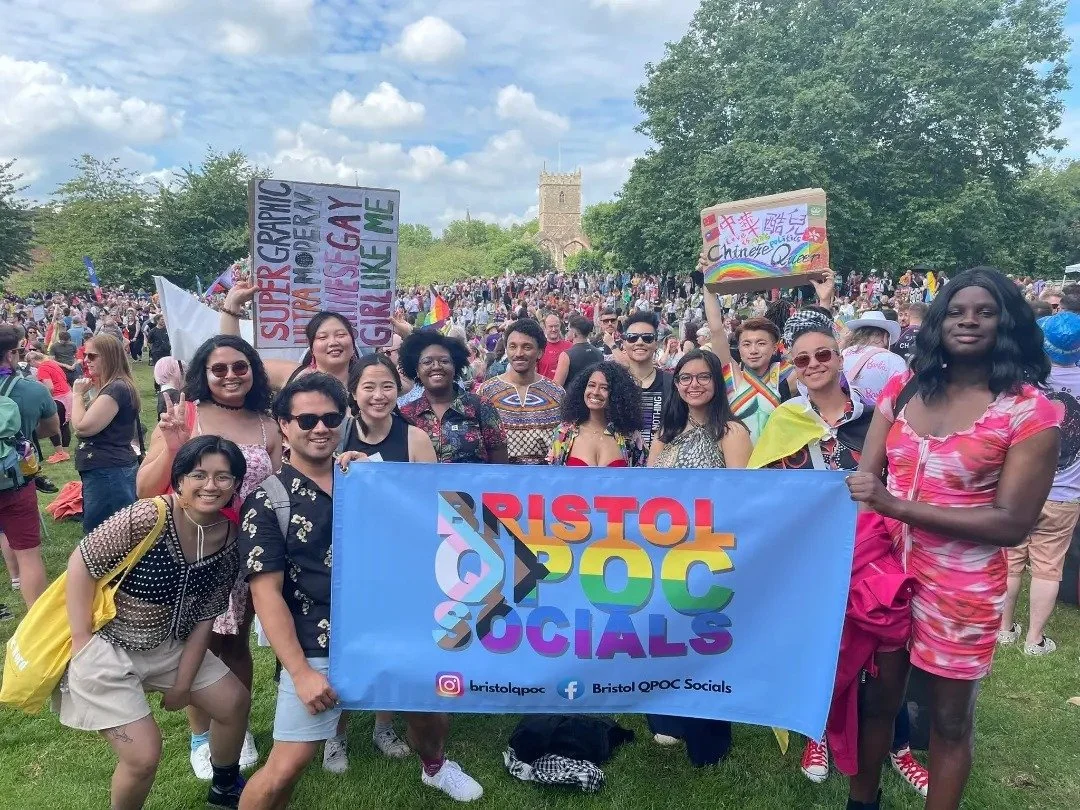 A diverse group of people holding a Bristol QPOC Socials banner at Bristol Pride outdoor event with many others in the background, some holding signs supporting LGBTQ+ pride, on a sunny day with green trees. In Castle Park with trans and queer banner
