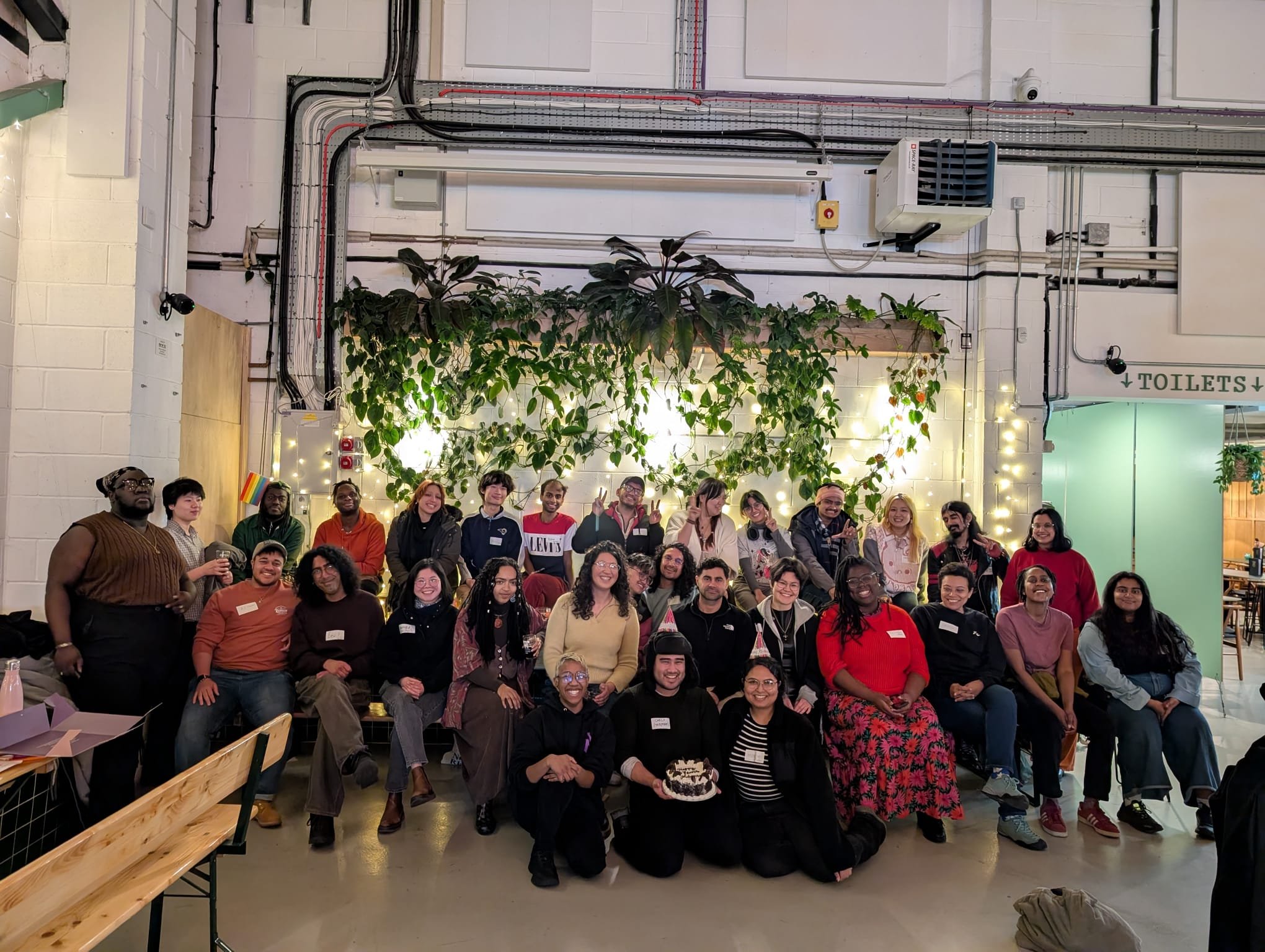 Group of diverse people gathered indoors, some wearing party hats, with a birthday cake, in front of a green leafy backdrop with string lights, celebrating together.