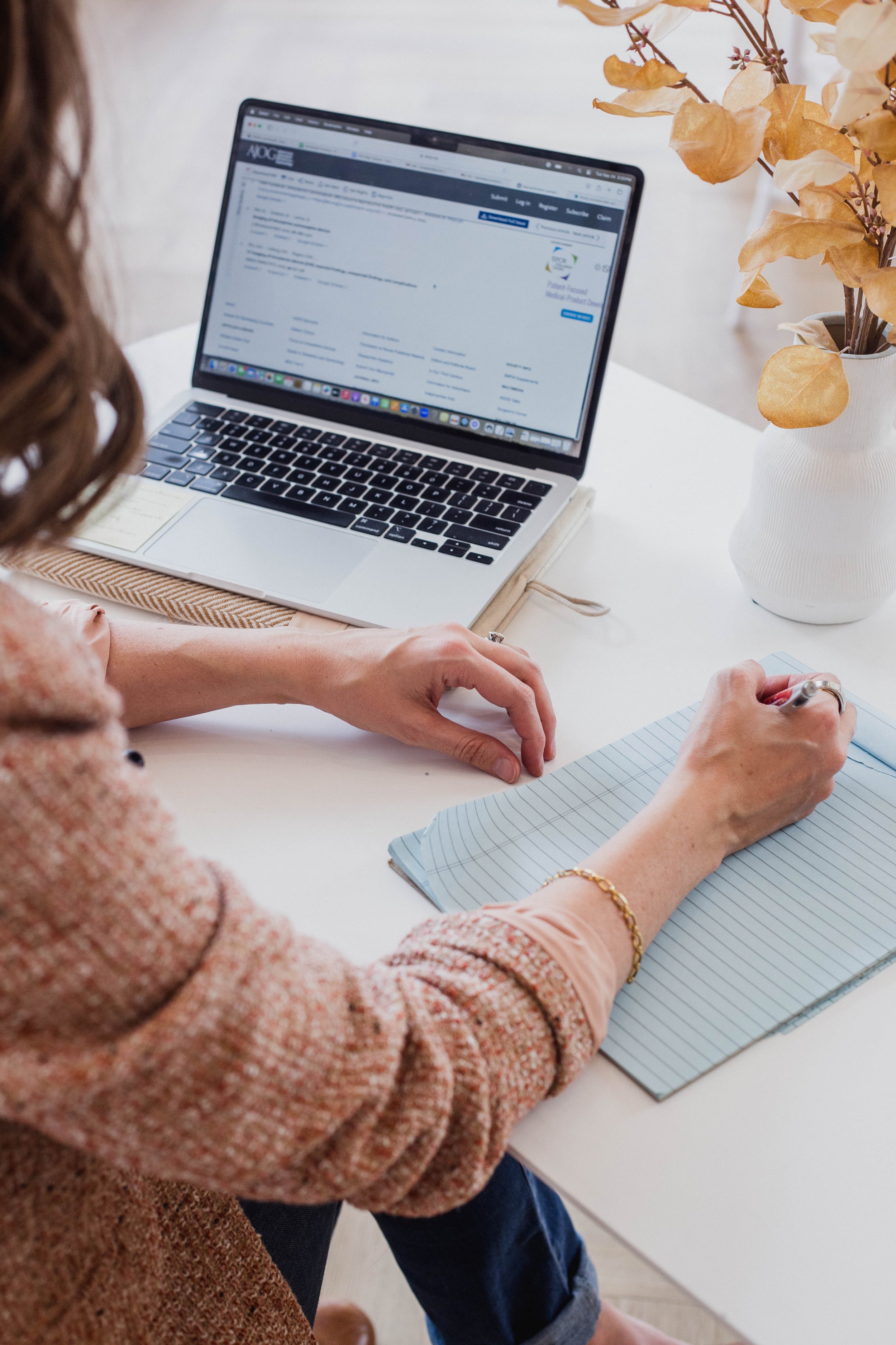 Person writing notes on a lined notepad at a white desk with a closed laptop and a white vase with dried flowers.