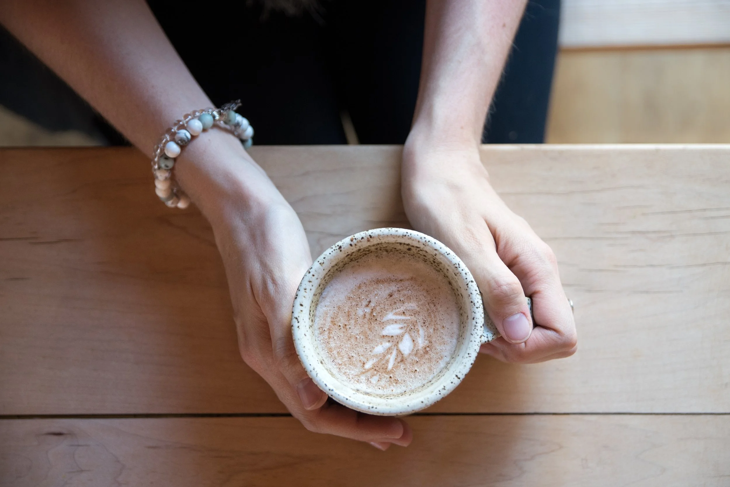 Person holding a ceramic mug of frothy coffee or latte with latte art on top, sitting at a wooden table, wearing a beaded bracelet.