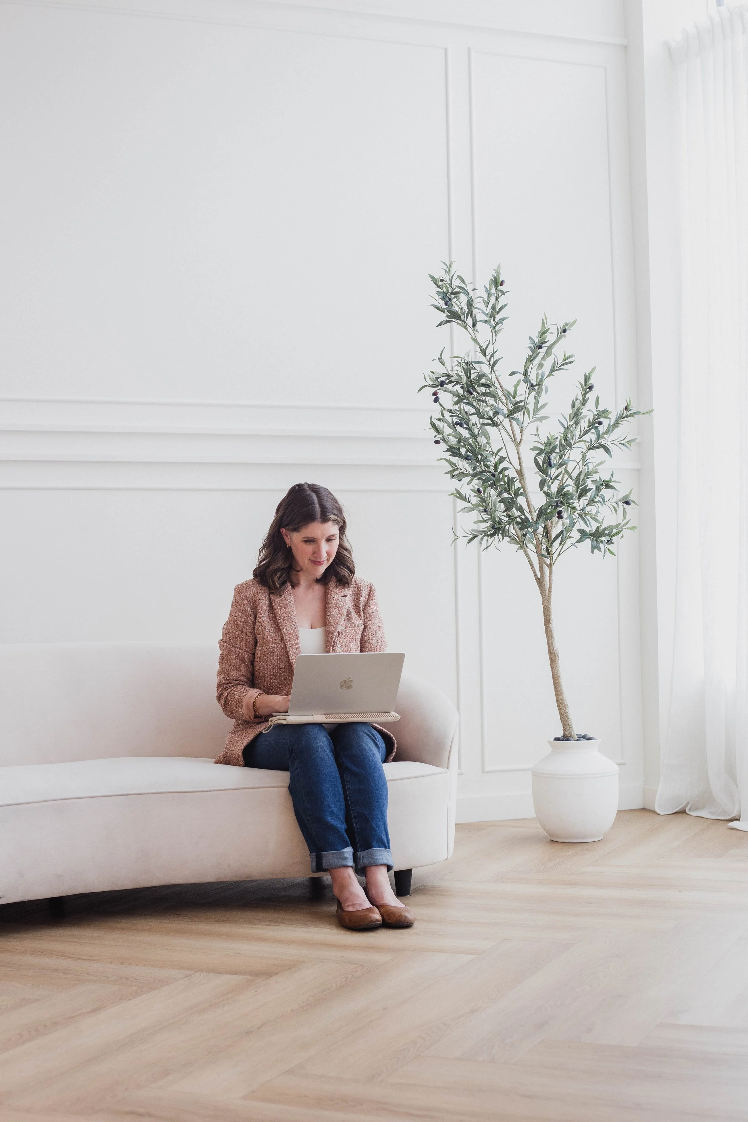 A woman sitting on a white couch with a laptop in a bright, minimalist room with white walls, wooden floor, and a large potted plant.