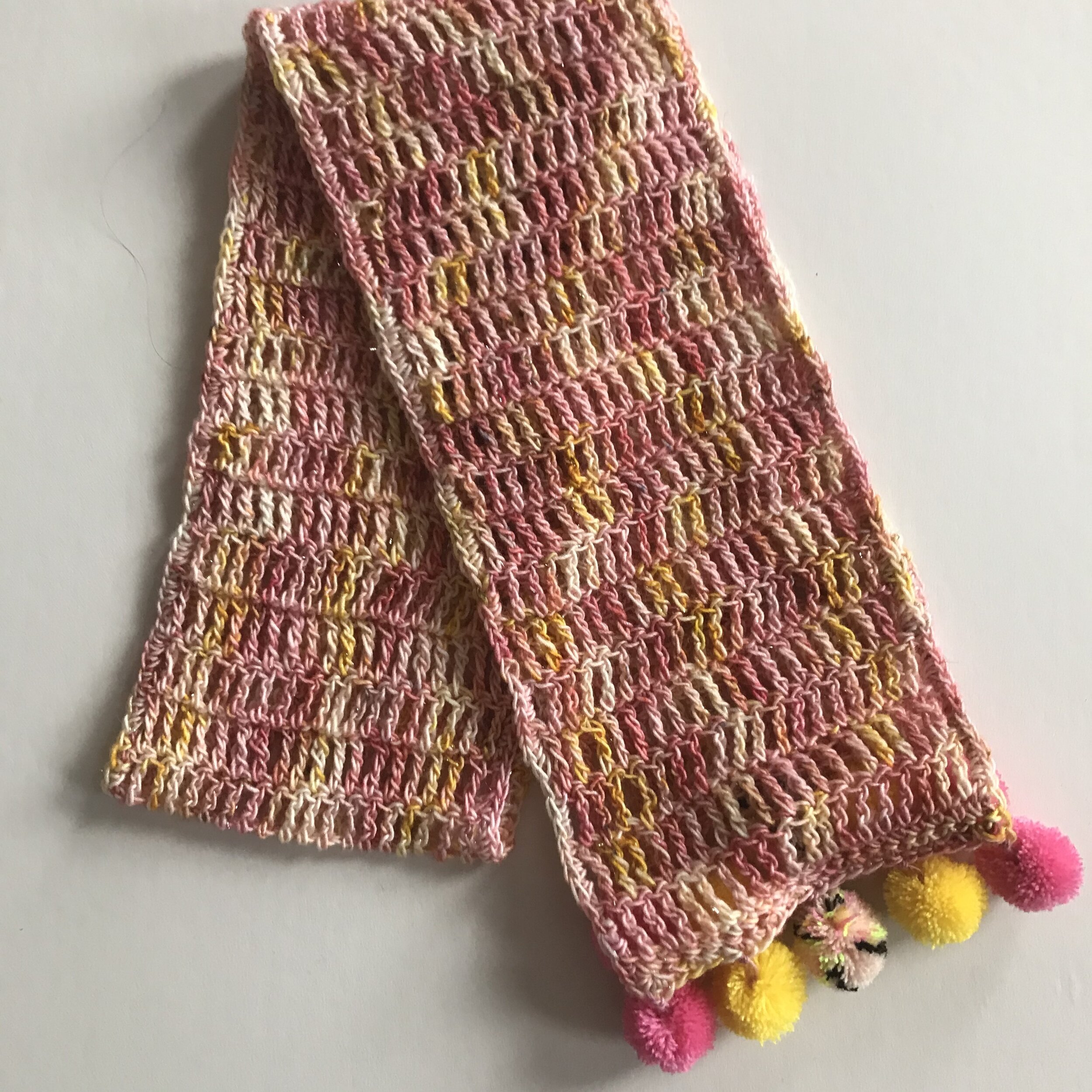 Colorful knitted scarf with pink, yellow, white, and beige yarn, decorated with pink, yellow, and multicolored pom-poms at the ends.