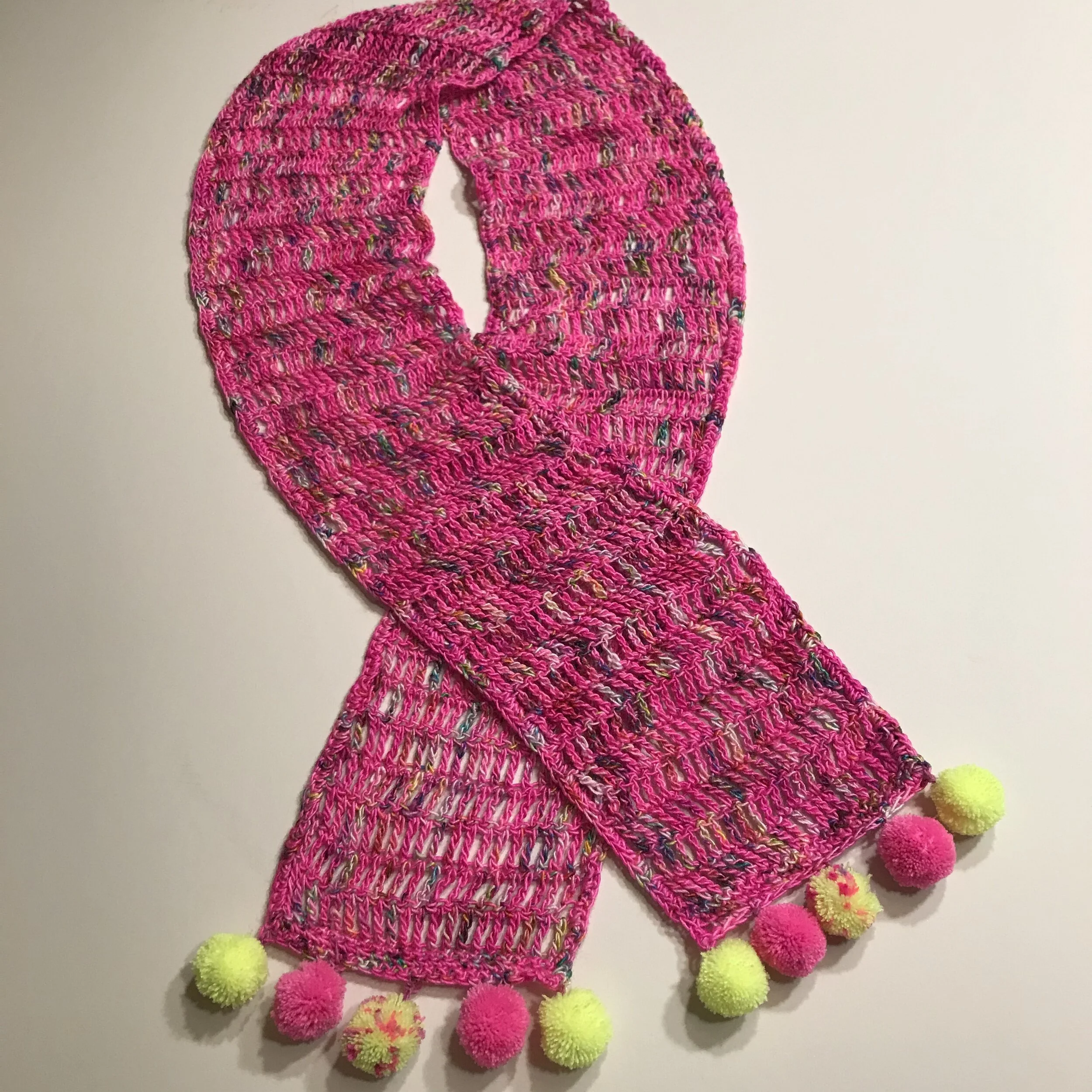 Pink knitted scarf with multicolored specks and yellow, pink, and multicolored pom-poms at the ends, laid flat on a white surface.