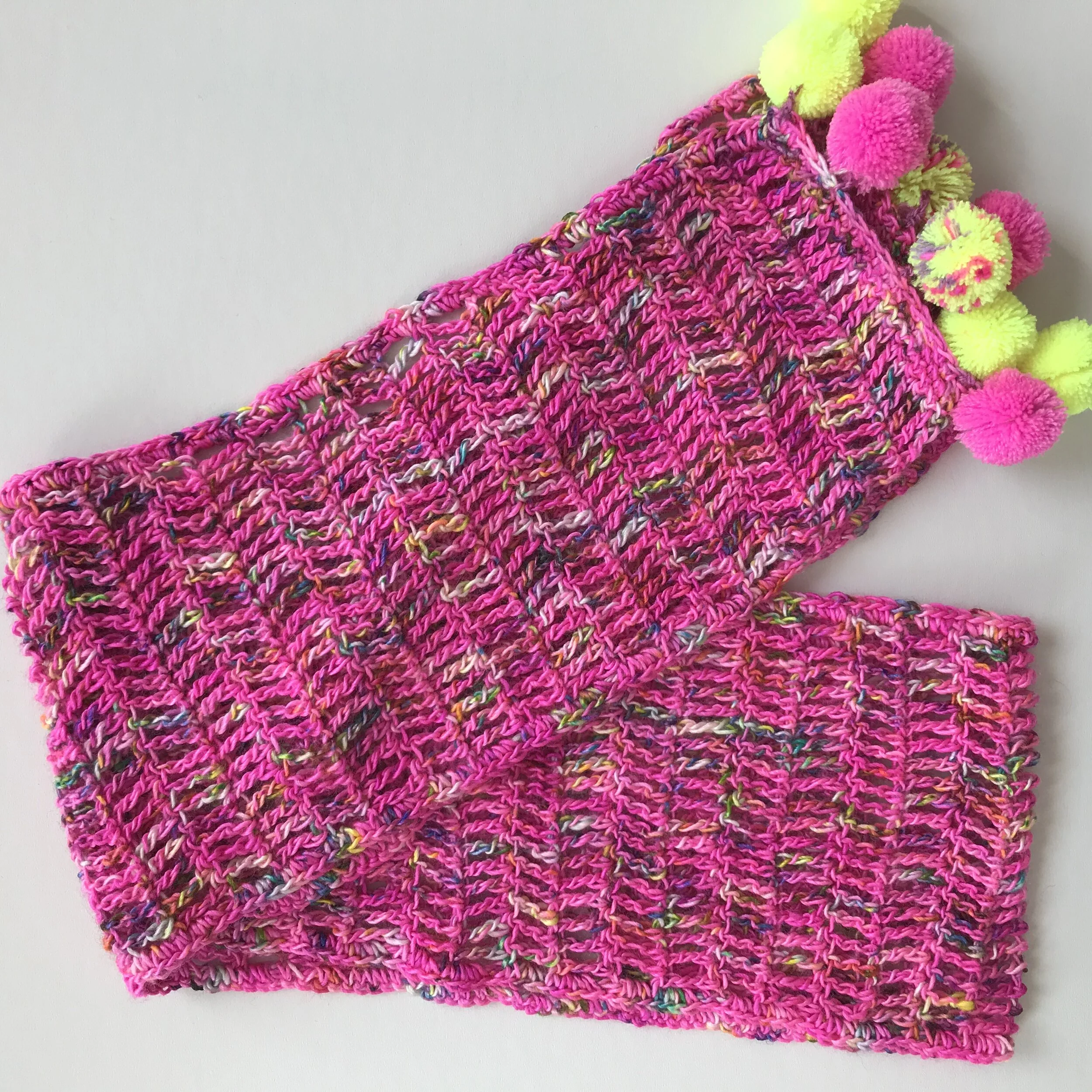 Pink knitted scarf with multicolored yarn and fuzzy pom-pom accents in pink and yellow.
