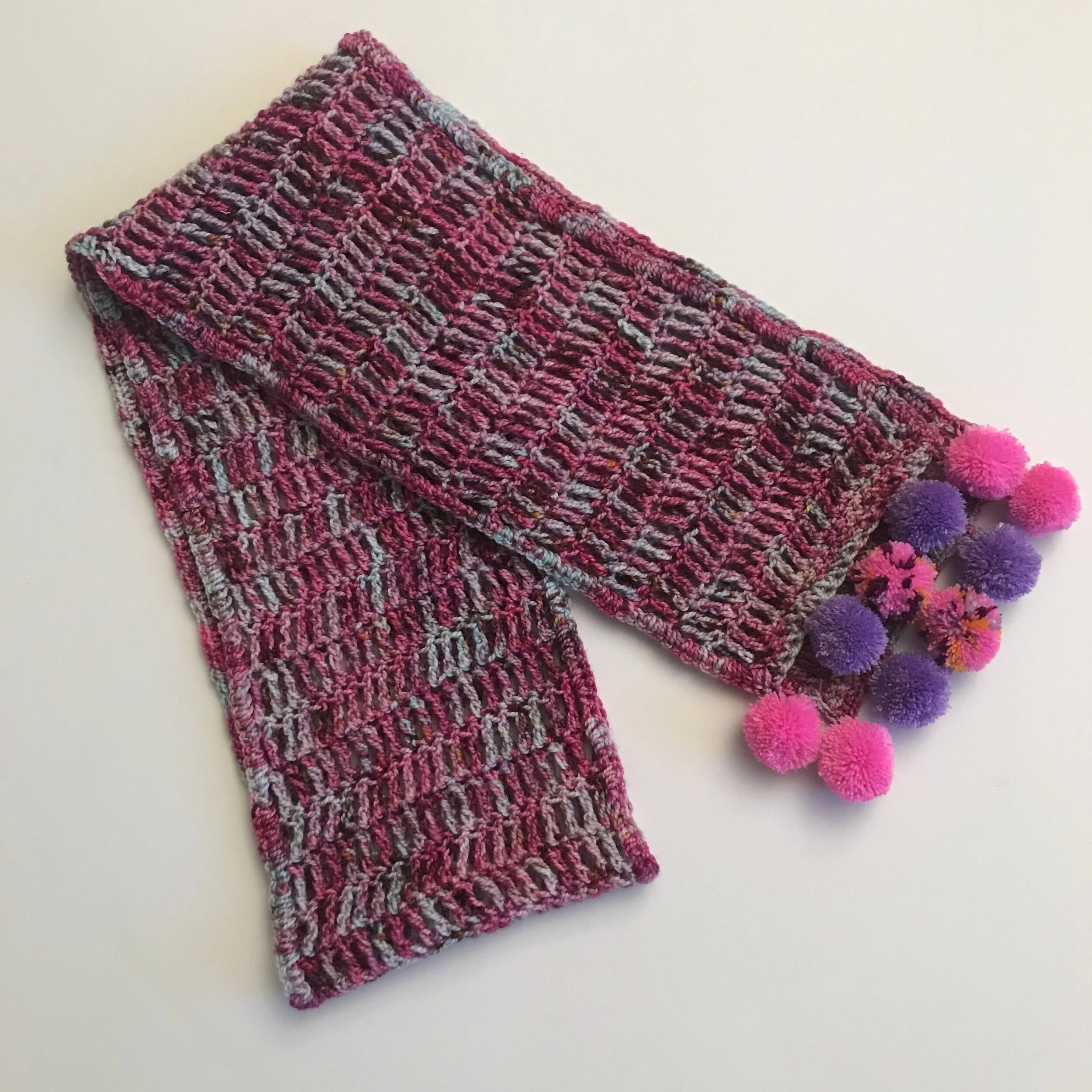 A multicolored knitted scarf with pink, purple, and gray tones, decorated with pink and purple pom-poms along one edge, laid out on a white surface.