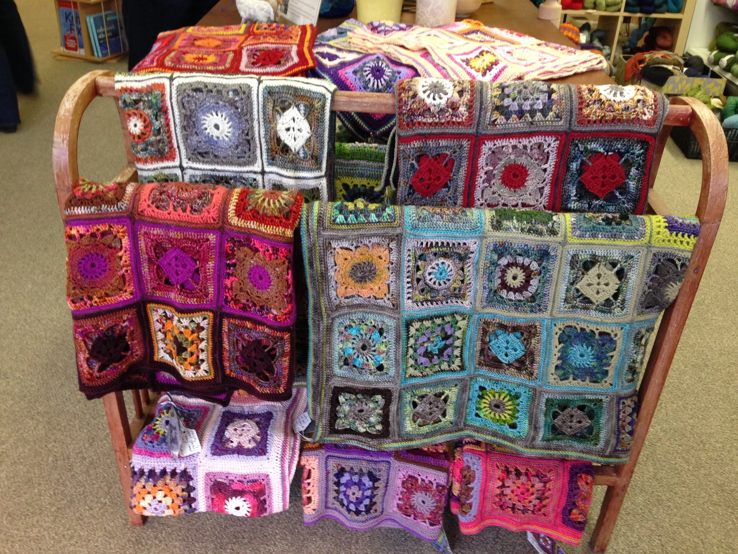 Colorful handmade crochet granny square blankets draped over a wooden display. Each blanket features various vibrant colors and intricate patterns.
