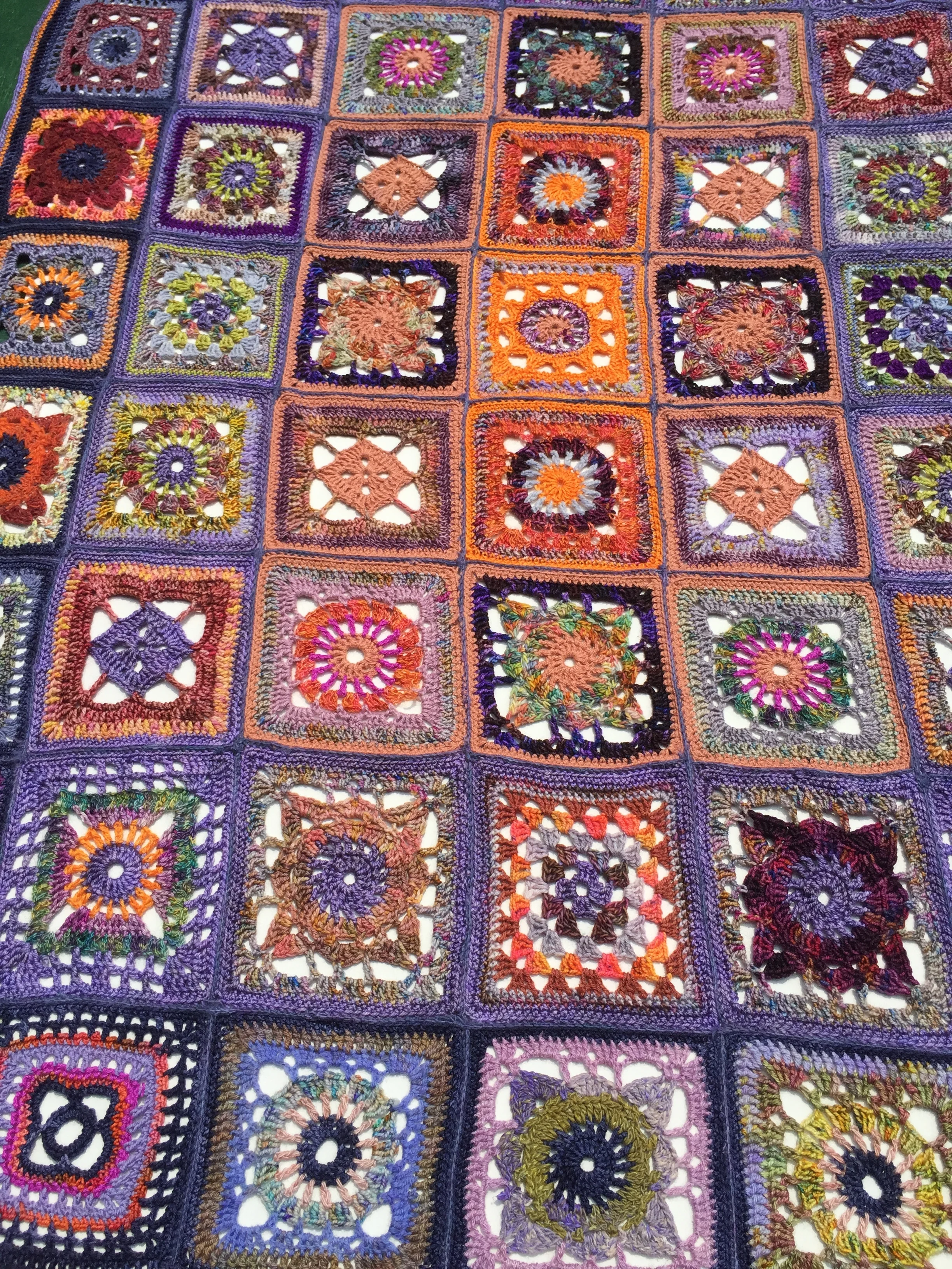 A colorful handmade crochet blanket composed of numerous square patches, each with intricate and diverse patterns, bordered with purple yarn.