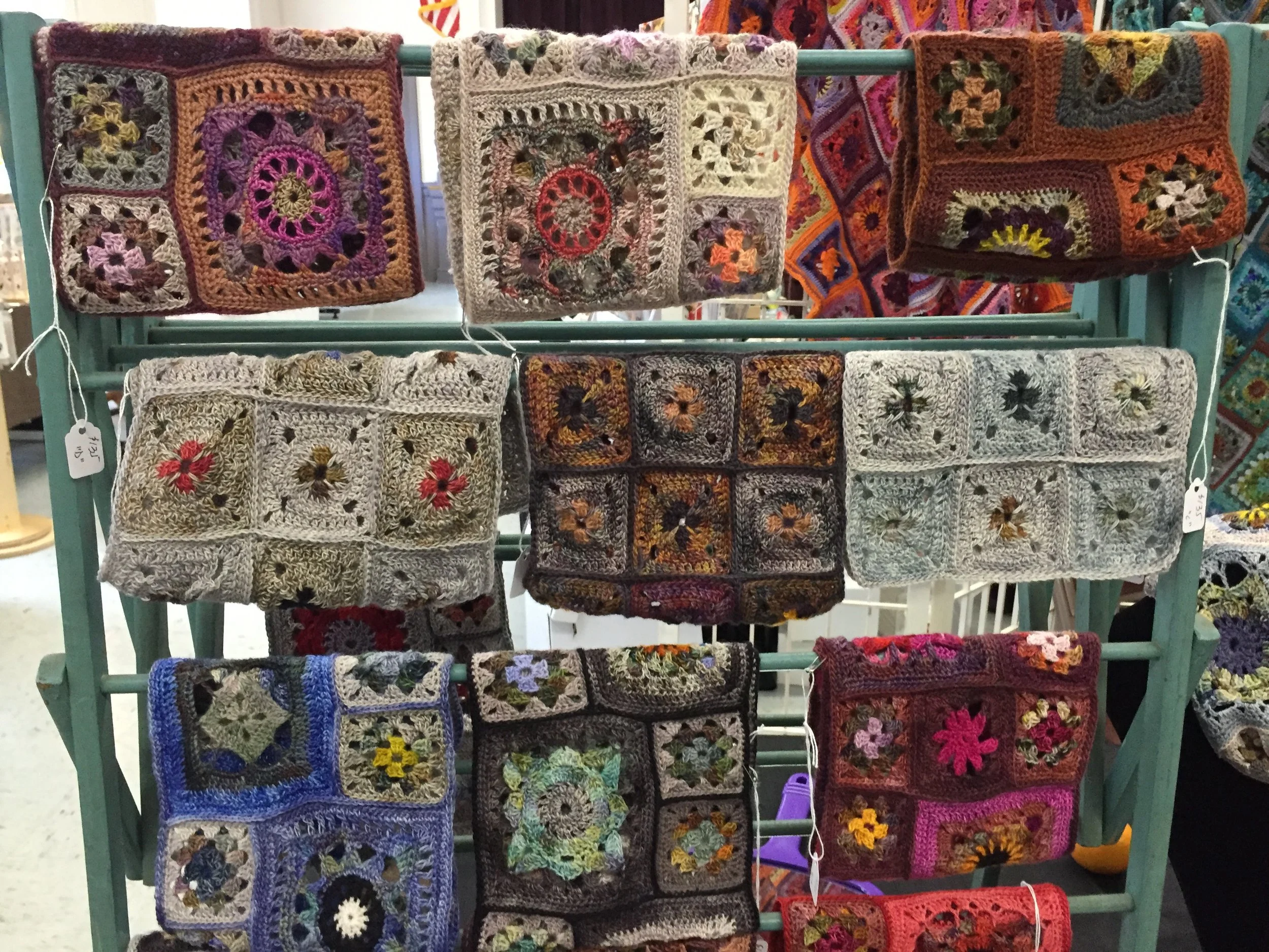 Display of handmade crochet granny square blankets hanging on a rack at a craft fair.