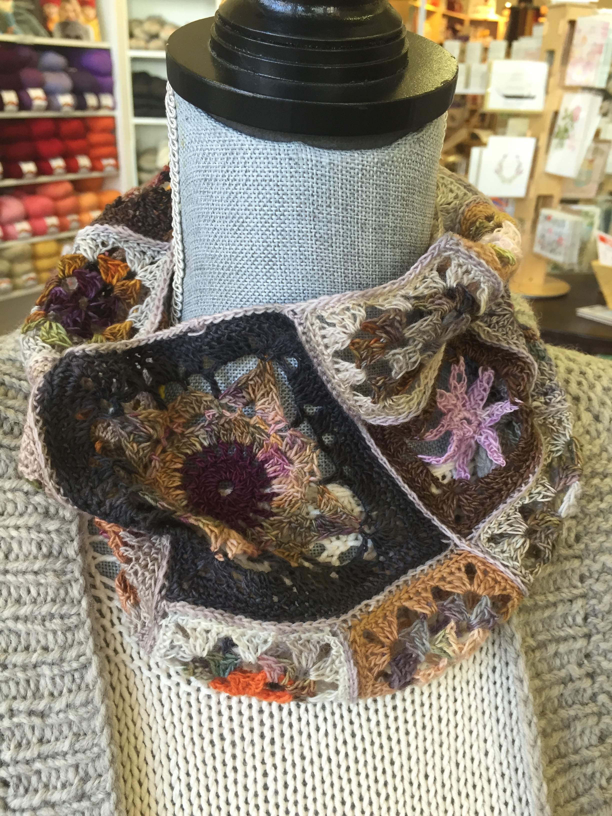 A mannequin displaying a colorful crocheted scarf featuring floral patterns with multiple colors including purple, pink, beige, and black, showcased against a background of yarns and craft supplies in a store.