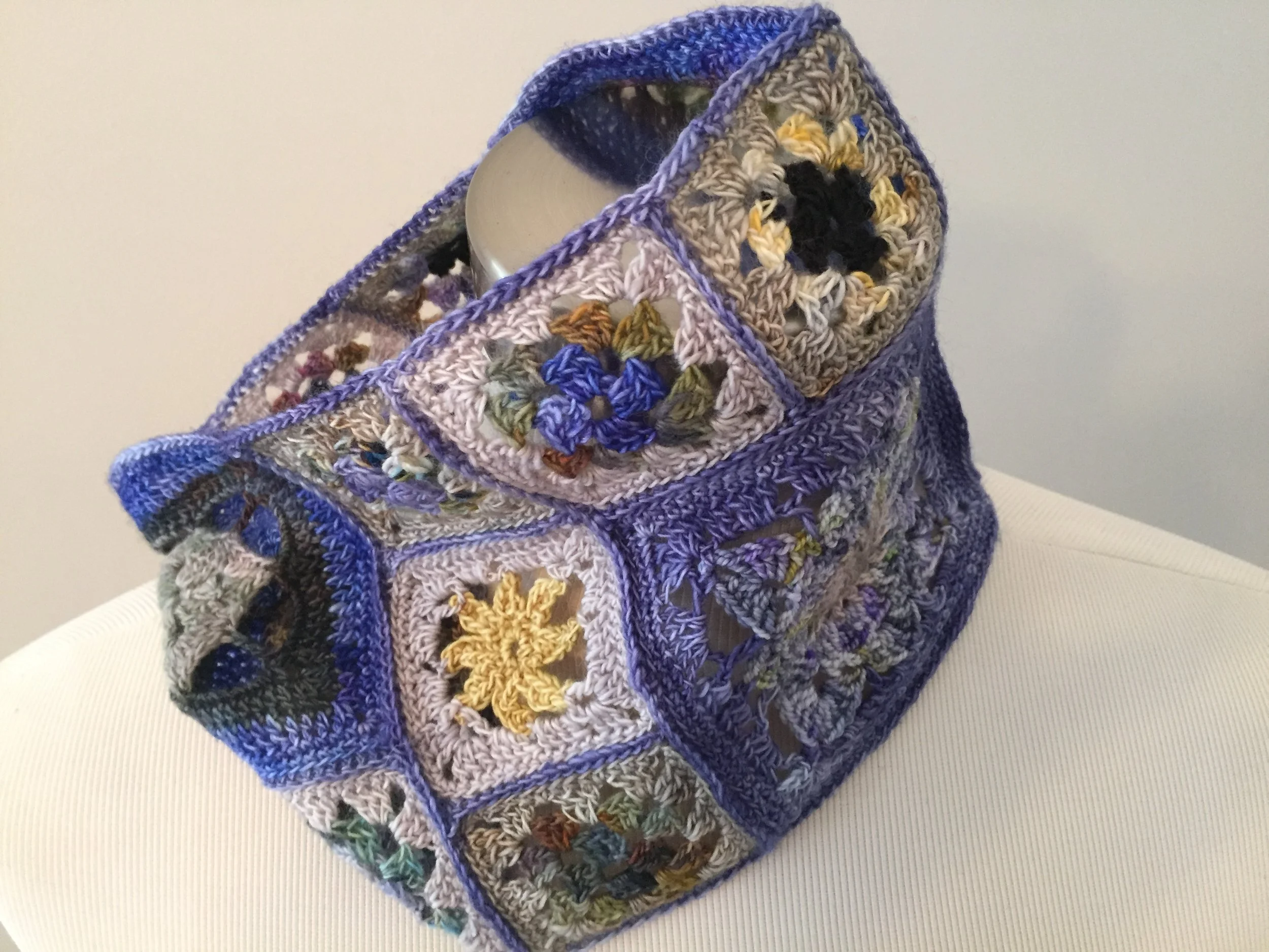 A colorful crocheted bag made of multiple granny squares with various colors and patterns, constructed with thick yarn, featuring a handle and a partial view of a cylindrical metallic object inside.
