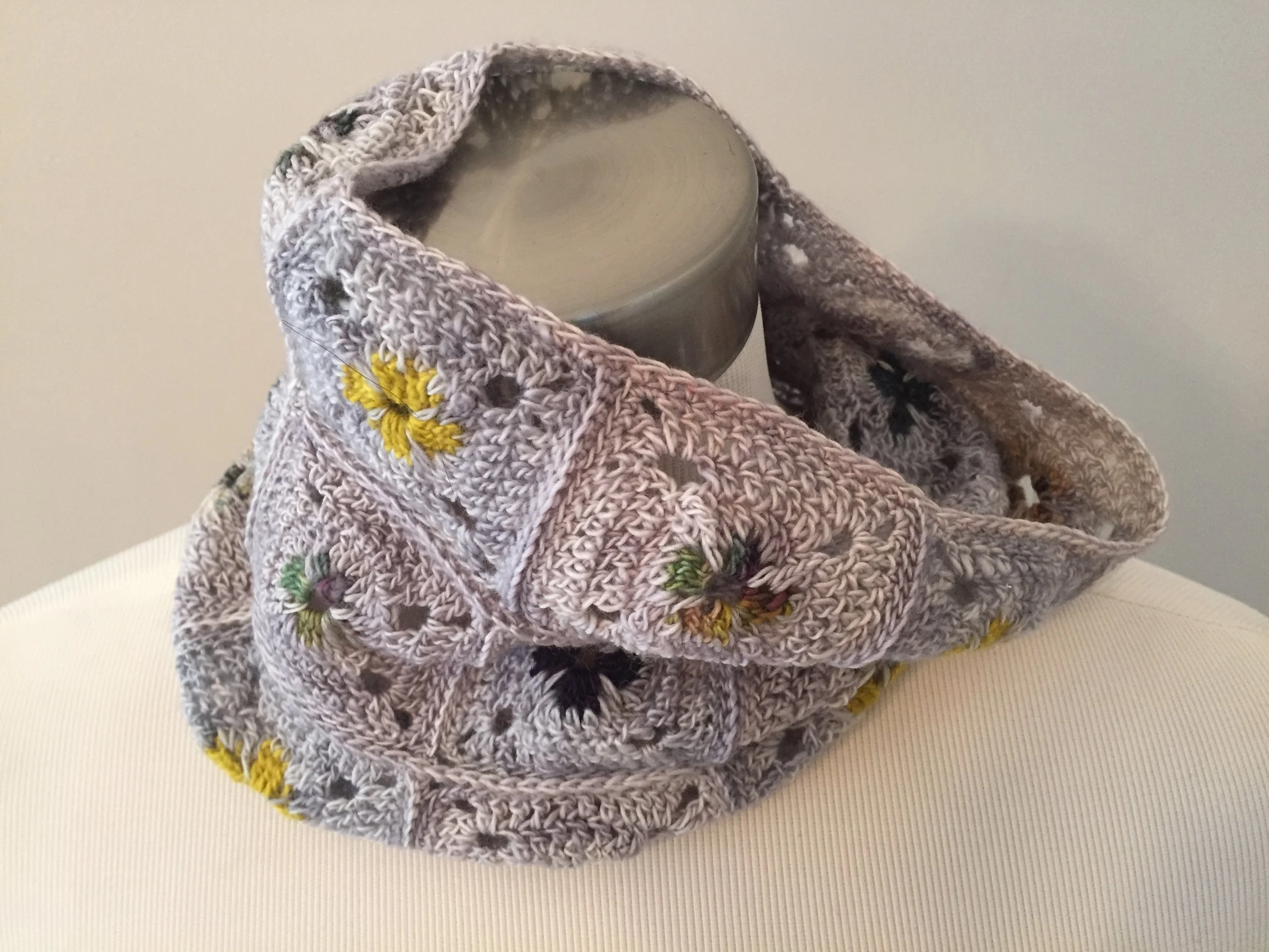 A grey knitted scarf with embroidered yellow, green, and black flowers, draped over a white surface with a ceramic mug partially placed under it.