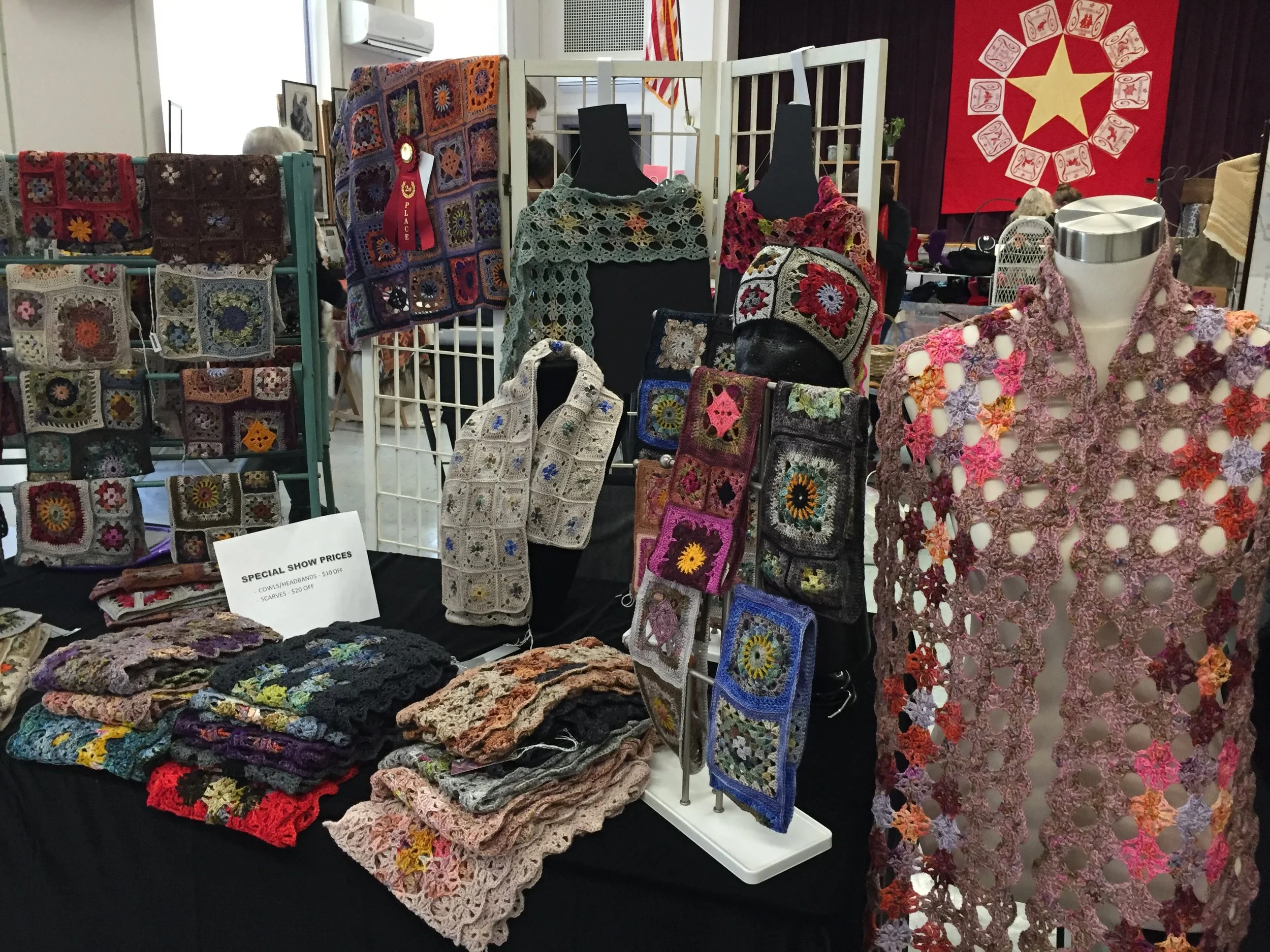 Display of handmade crochet scarves, shawls, and accessories at a craft fair booth.