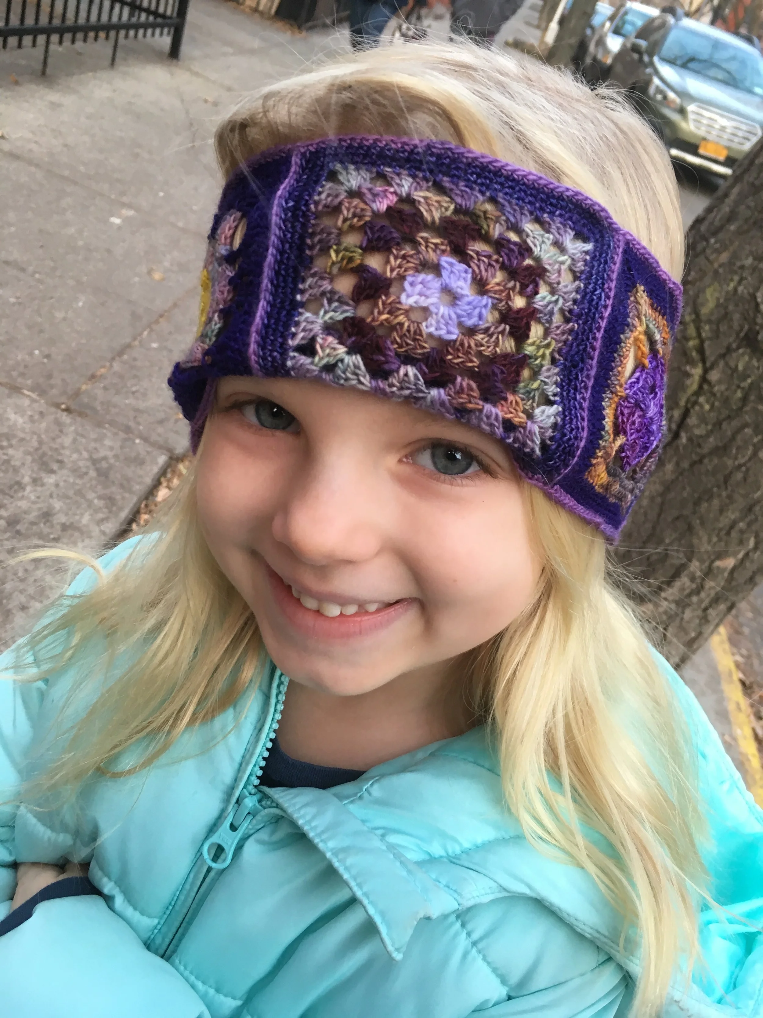 A young girl outdoors wearing a purple crochet headband with floral patterns and a light blue puffer jacket, smiling at the camera.