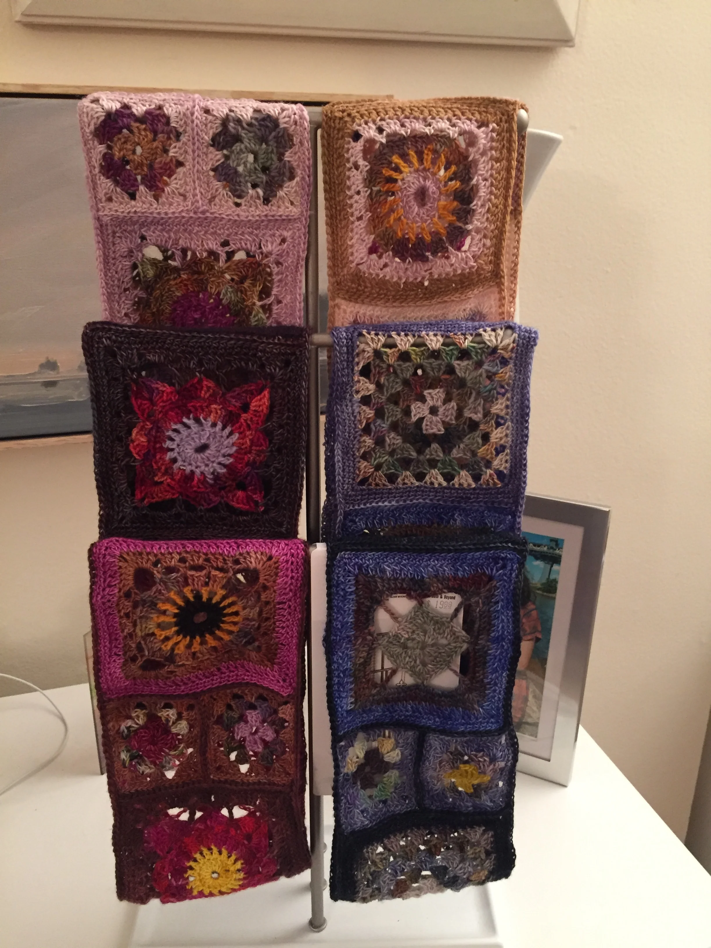 A white display stand holding ten small handmade crocheted square items, each with colorful floral or geometric patterns. The stand is on a white surface with a framed photo in the background.