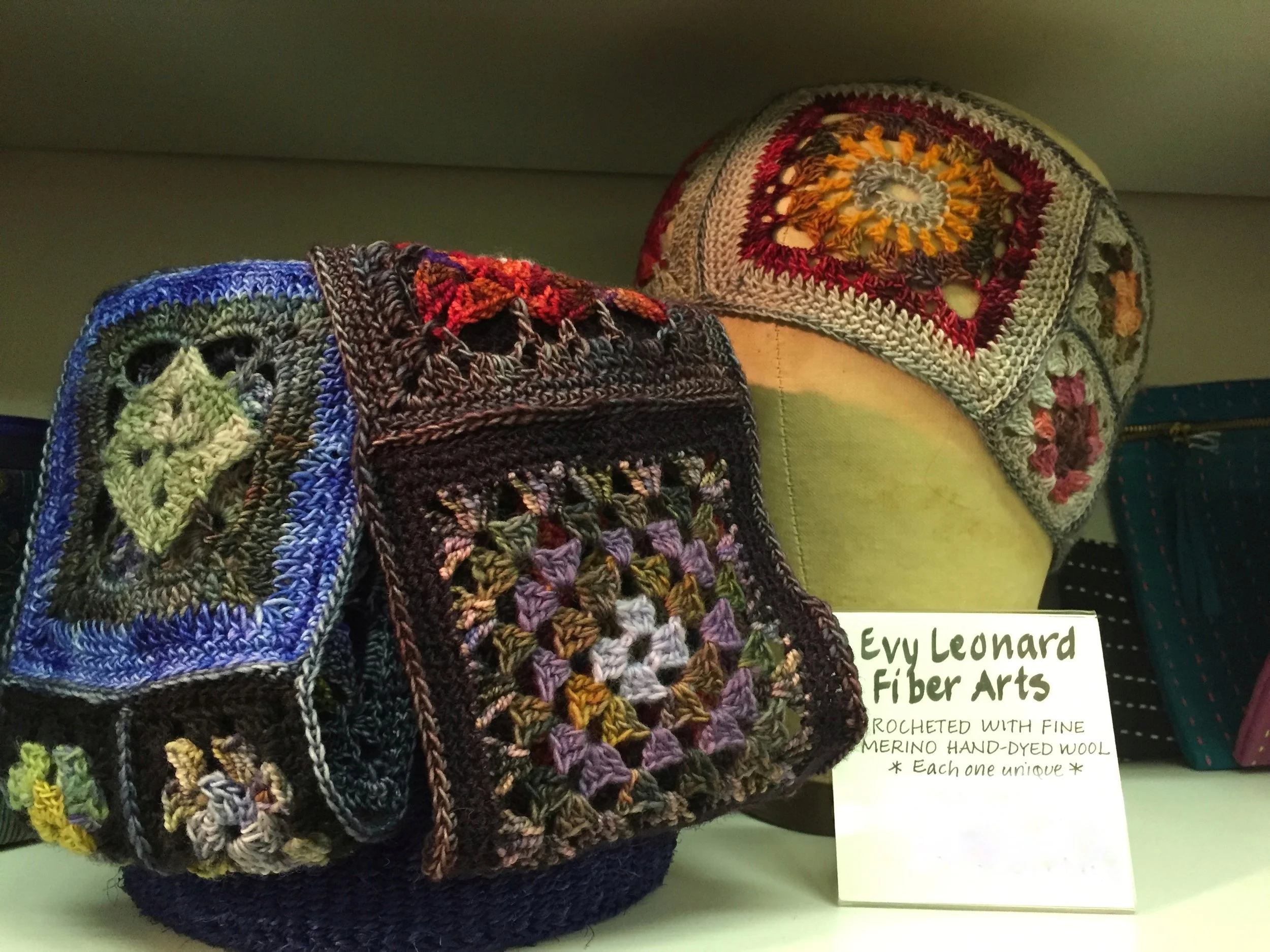 Display of hand-crafted crocheted bags with multicolored granny square patterns, text sign reads 'Evy Leonard Fiber Arts, crocheted with fine merino hand-dyed wool, each one unique'.