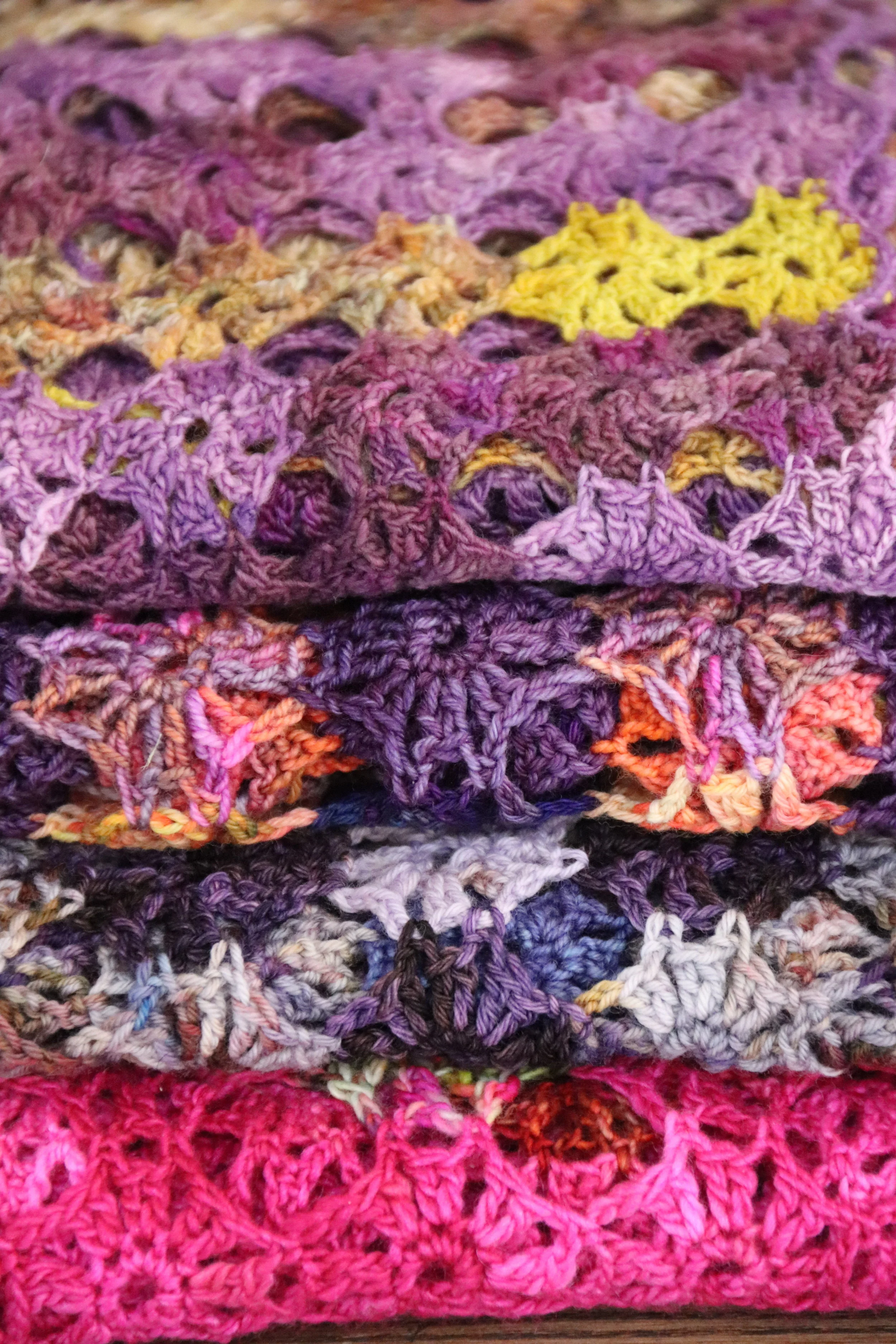 Stacked multicolored crochet scarves or blankets in purple, pink, gray, and yellow tones.