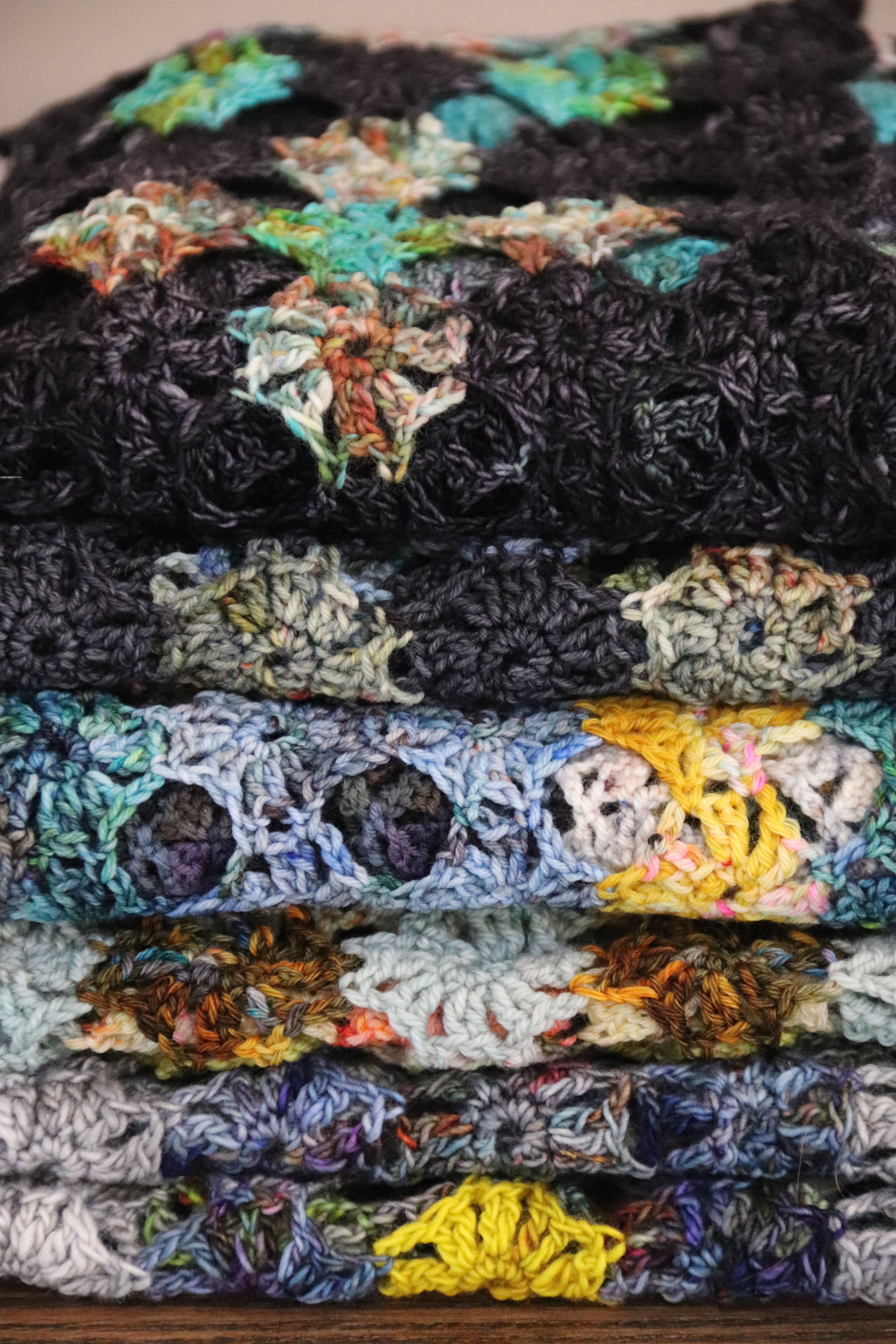 Stack of multicolored, crocheted blankets or throws, featuring various patterns and yarn textures.