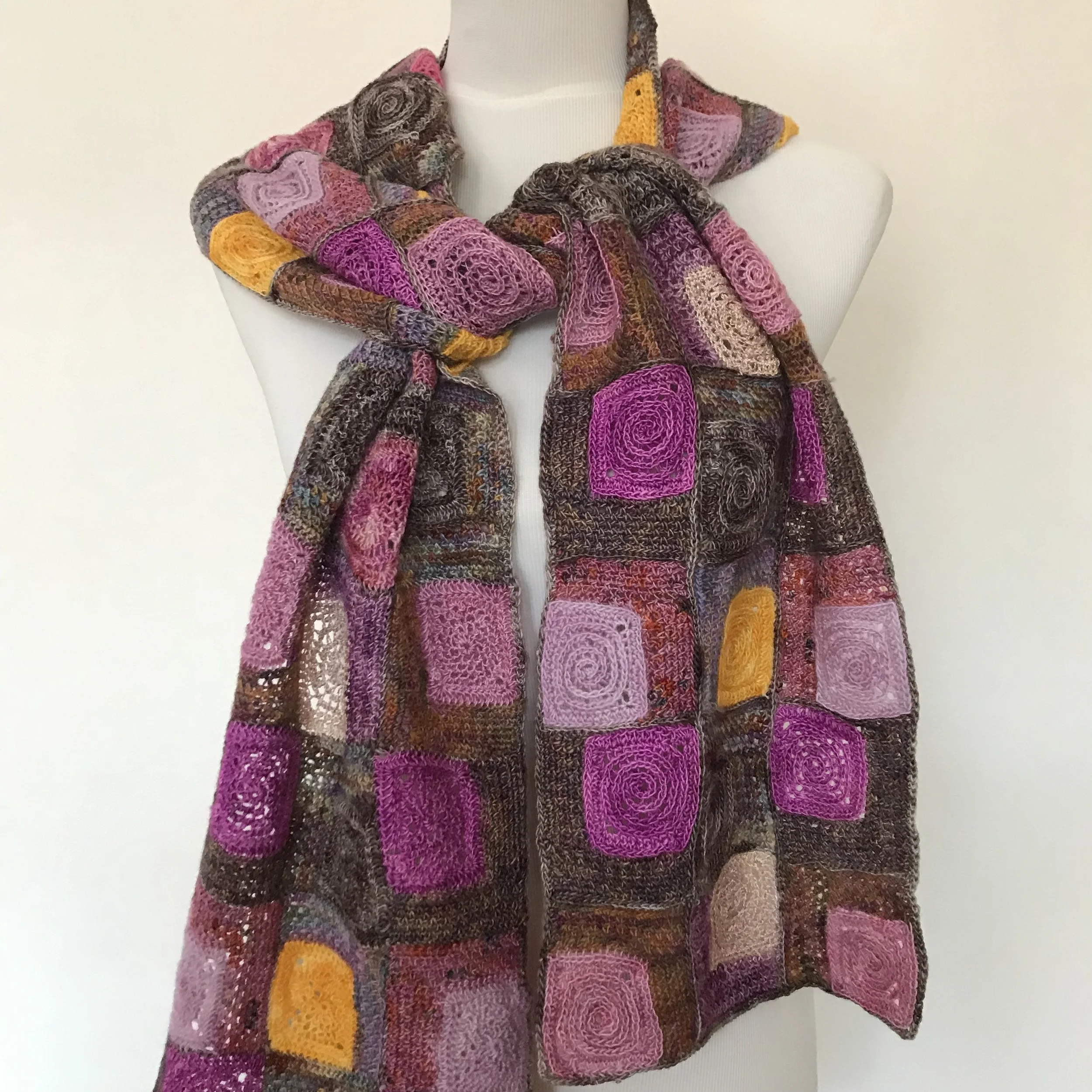 Colorful hand-knit patchwork scarf with various square patterns in shades of pink, purple, orange, and brown draped over a white mannequin.