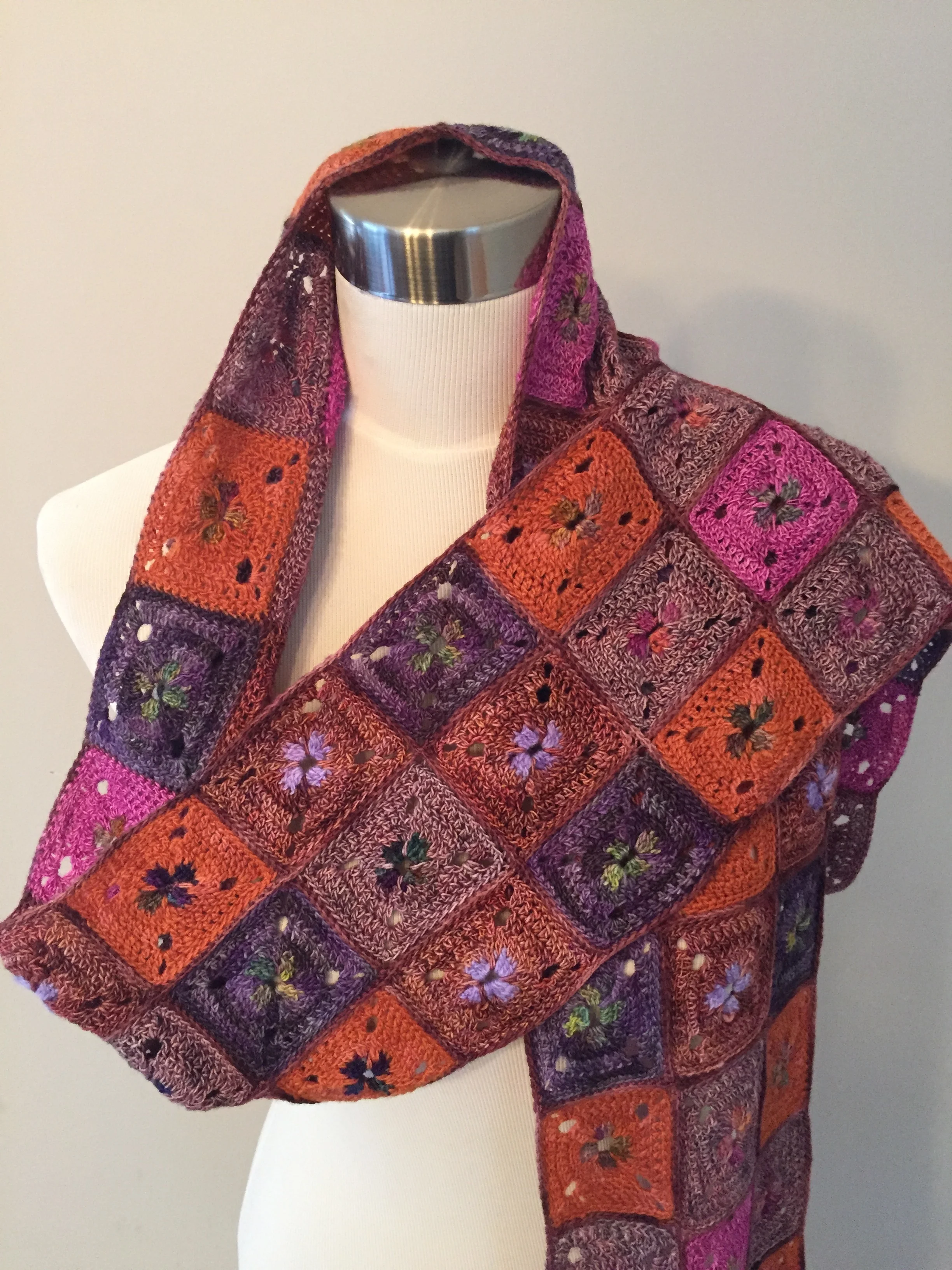Colorful knitted shawl with floral and geometric patterns draped over a mannequin with a white turtleneck, positioned against a plain light gray background.