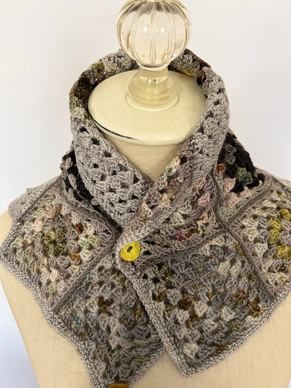 Close-up of a mannequin displaying a crocheted scarf with a mix of gray, black, pink, and green yarns and a yellow button.