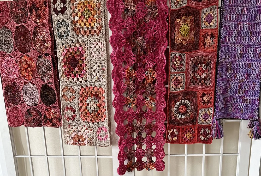Colorful crochet and knitting scarves and shawls hanging on a white rack.