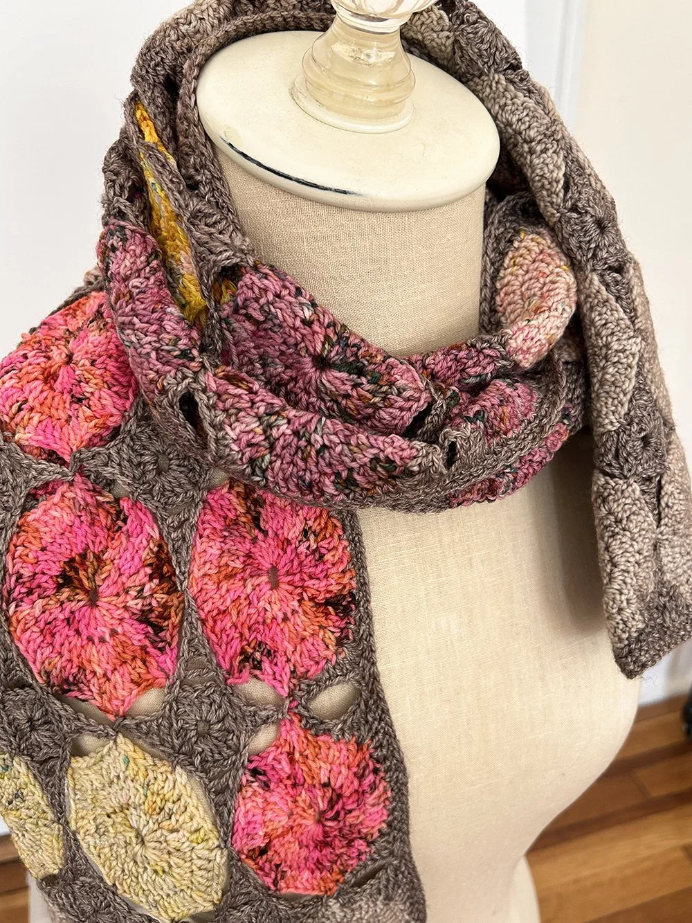 A crochet scarf with a floral pattern featuring pink, yellow, and multicolored flowers on a gray background, draped on a cream-colored mannequin bust.