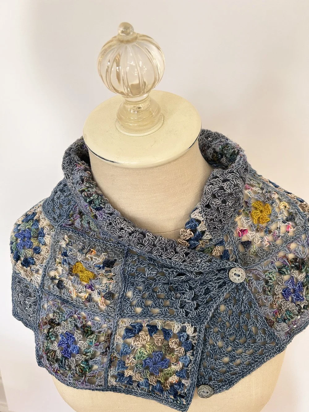 A mannequin dressed in a multicolored crocheted shawl with floral patterns, button fastenings, and a decorative glass finial on top.