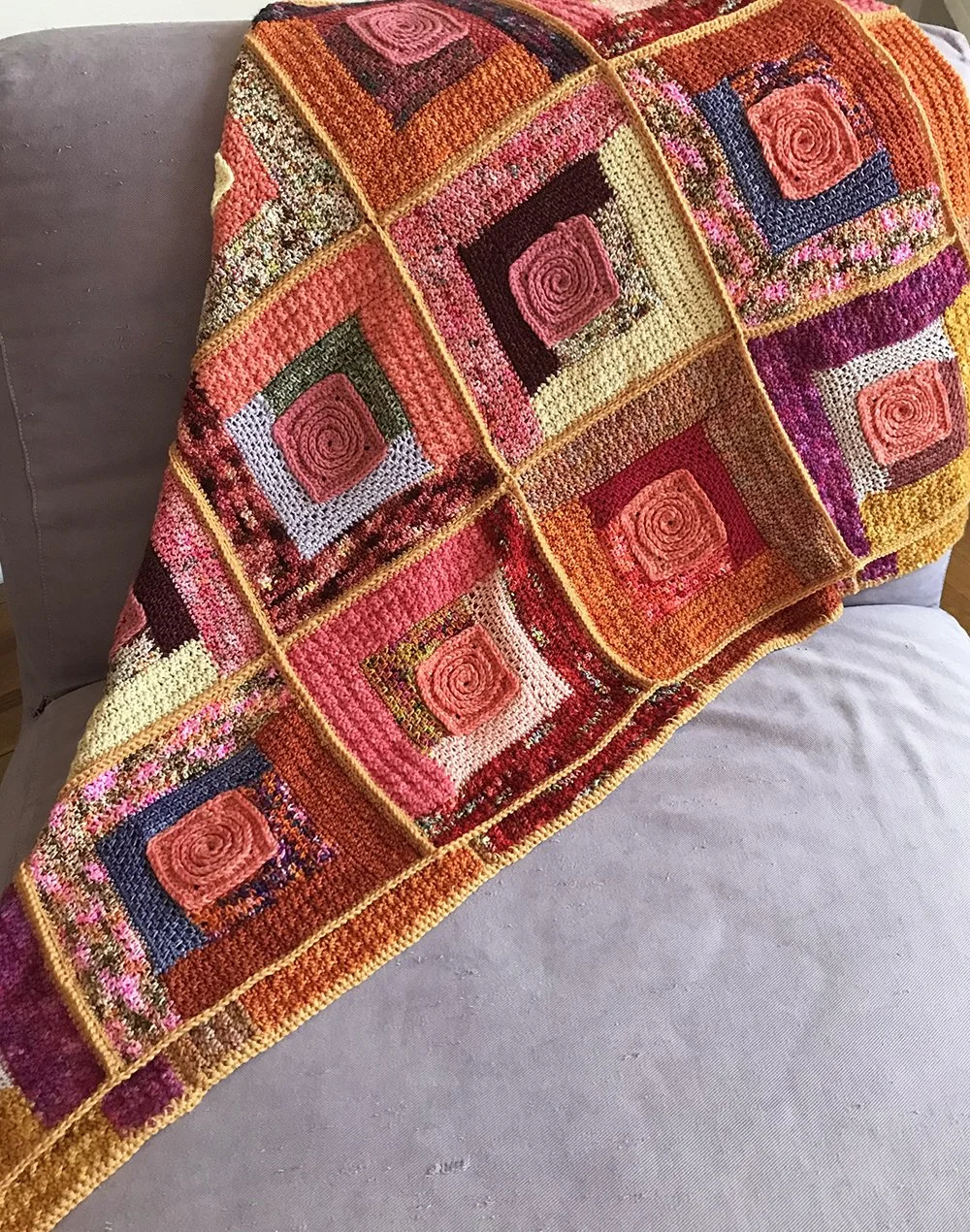 A colorful crocheted blanket with a patchwork design of square motifs in pink, red, orange, purple, and yellow, draped over a gray couch.