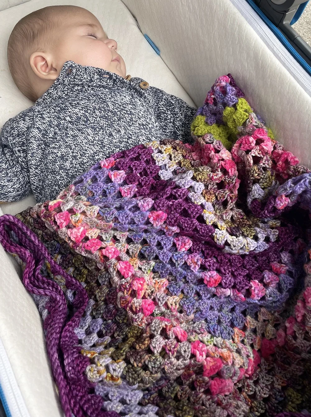 A sleeping baby lying in a bed covered with a colorful knitted blanket.