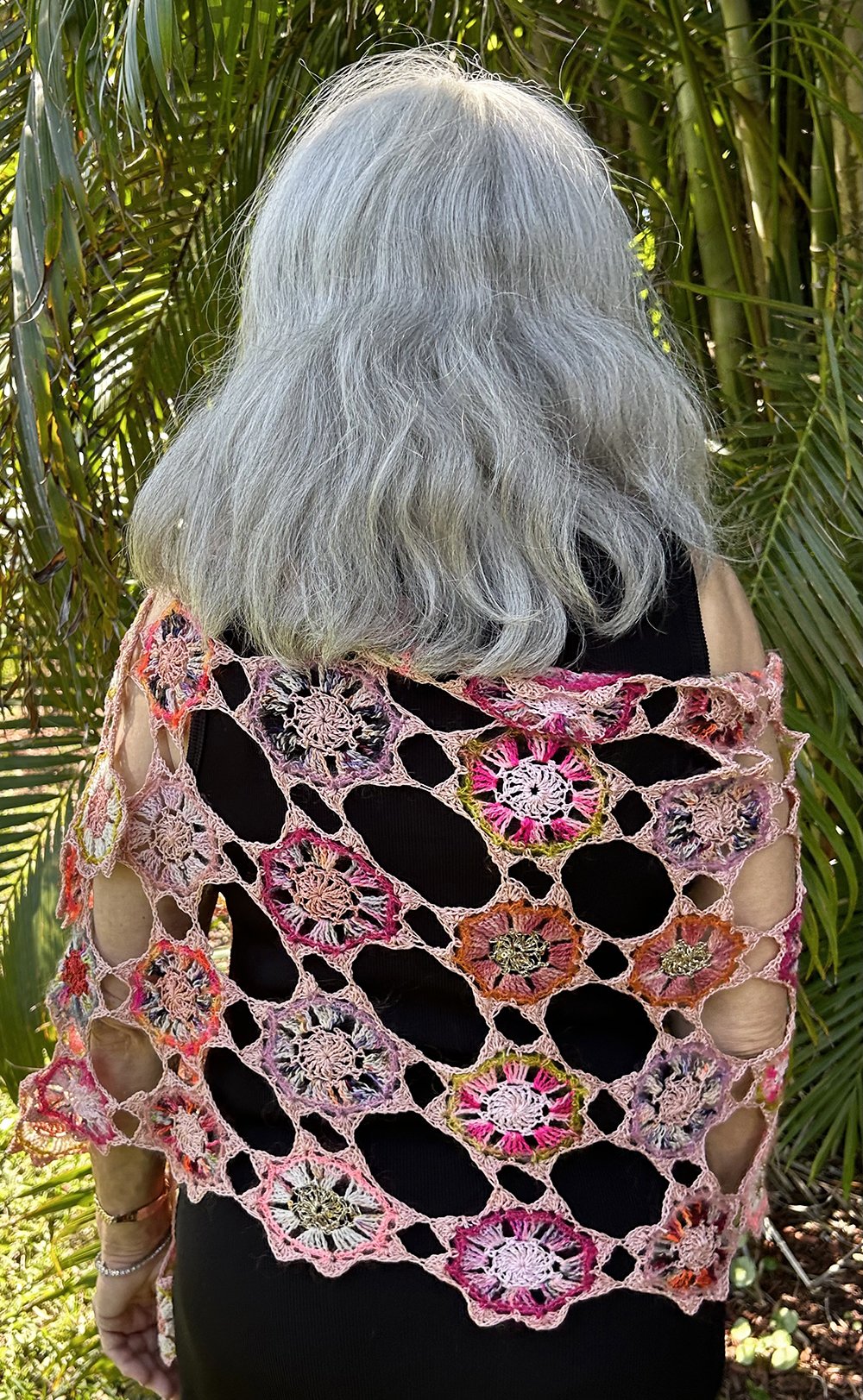 Back view of a person with long gray hair wearing a handmade, colorful crochet shawl featuring floral patterns, standing among green tropical plants.
