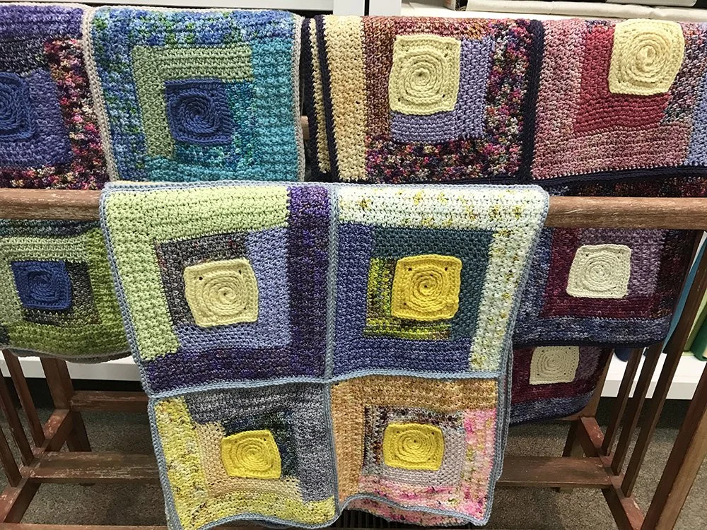 Multiple colorful knitted or crocheted blankets with spiral square patterns draped over a wooden rack.