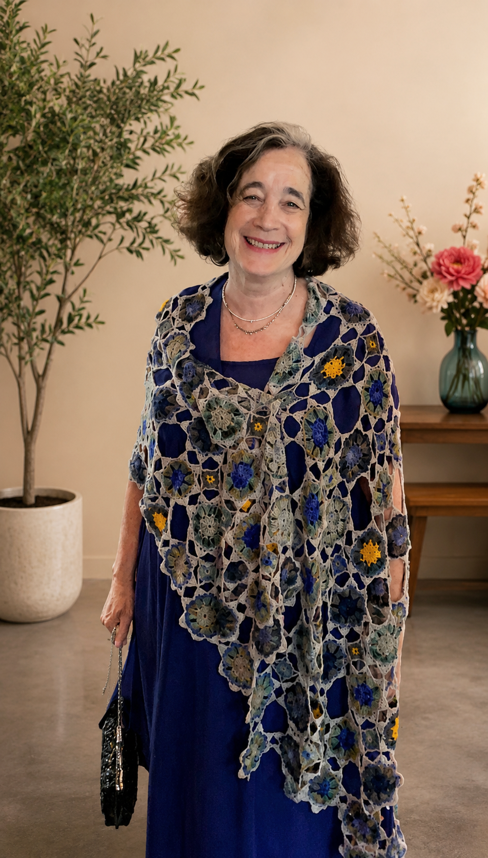 A woman standing indoors wearing a blue dress with a floral crochet shawl, smiling, with a potted plant and a vase with flowers in the background.