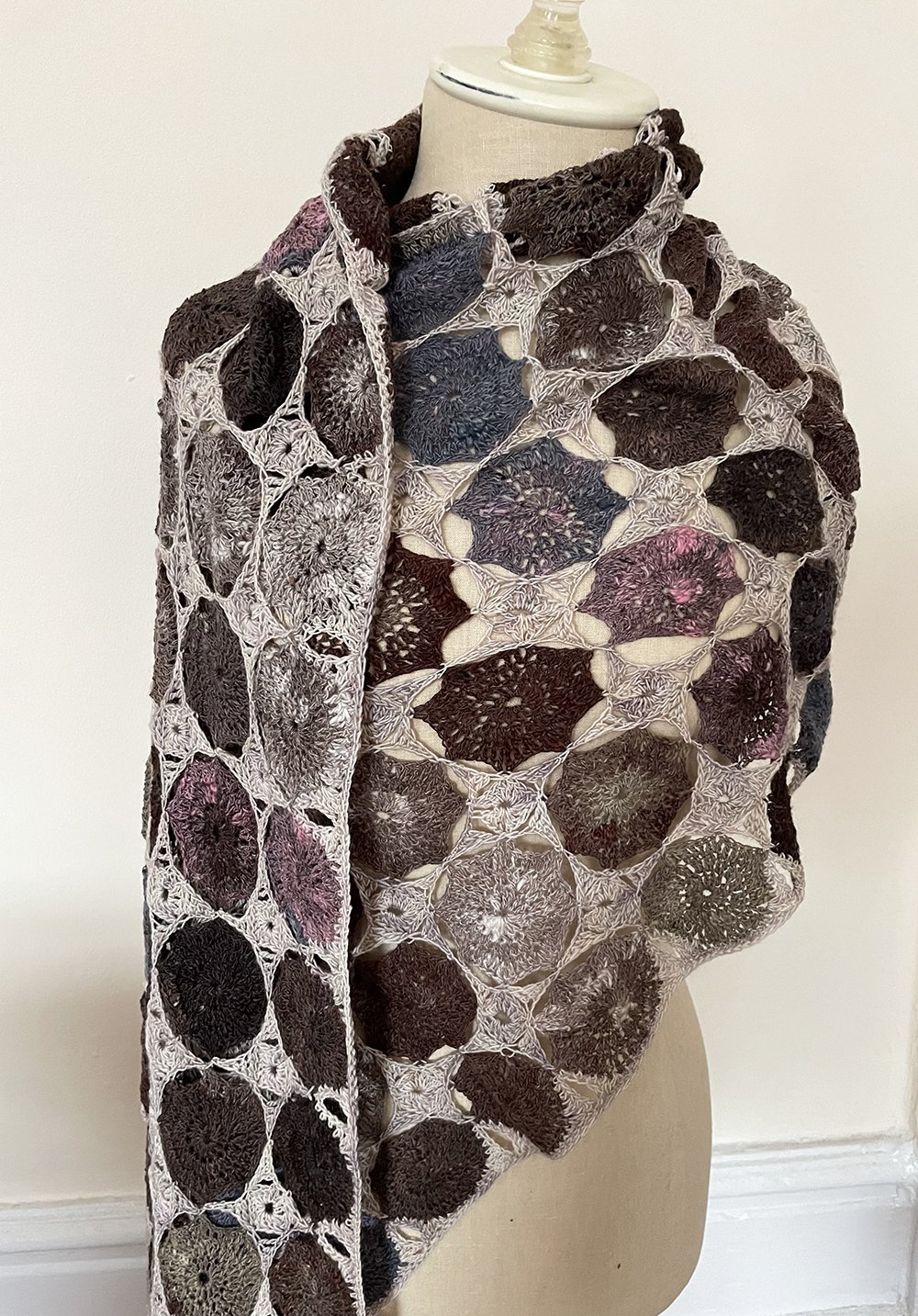A mannequin draped with a multicolored crochet shawl featuring hexagonal patterns in earth tones and shades of purple and pink.