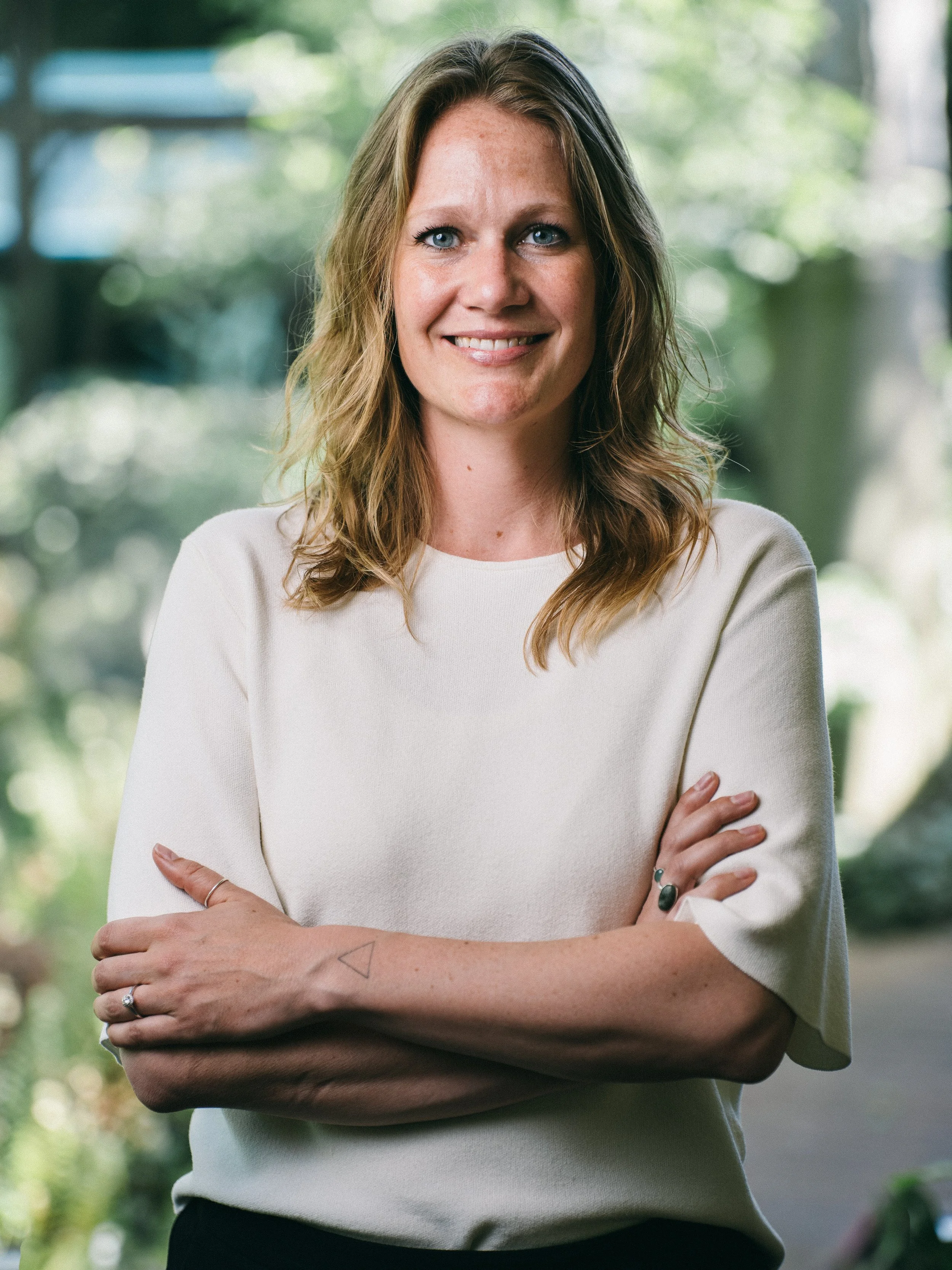 Porträt einer lachenden Frau mit blonden, gewellten Haaren, die vor einem grünen, bewaldeten Hintergrund steht, mit verschränkten Armen und einem weißen Oberteil.
