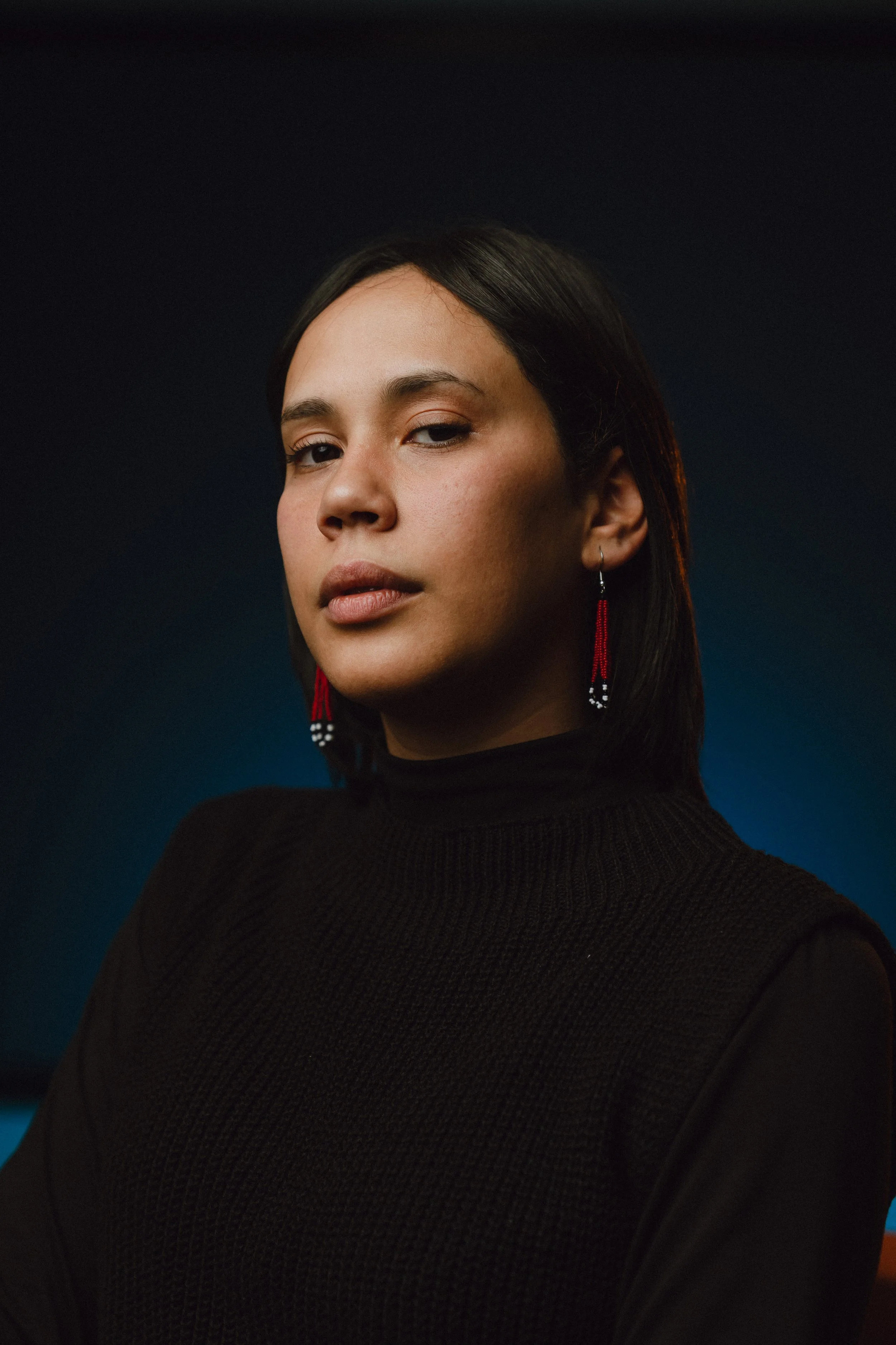 A woman with dark hair, wearing a black turtleneck sweater and red beaded earrings, posing with a serious expression against a dark background.