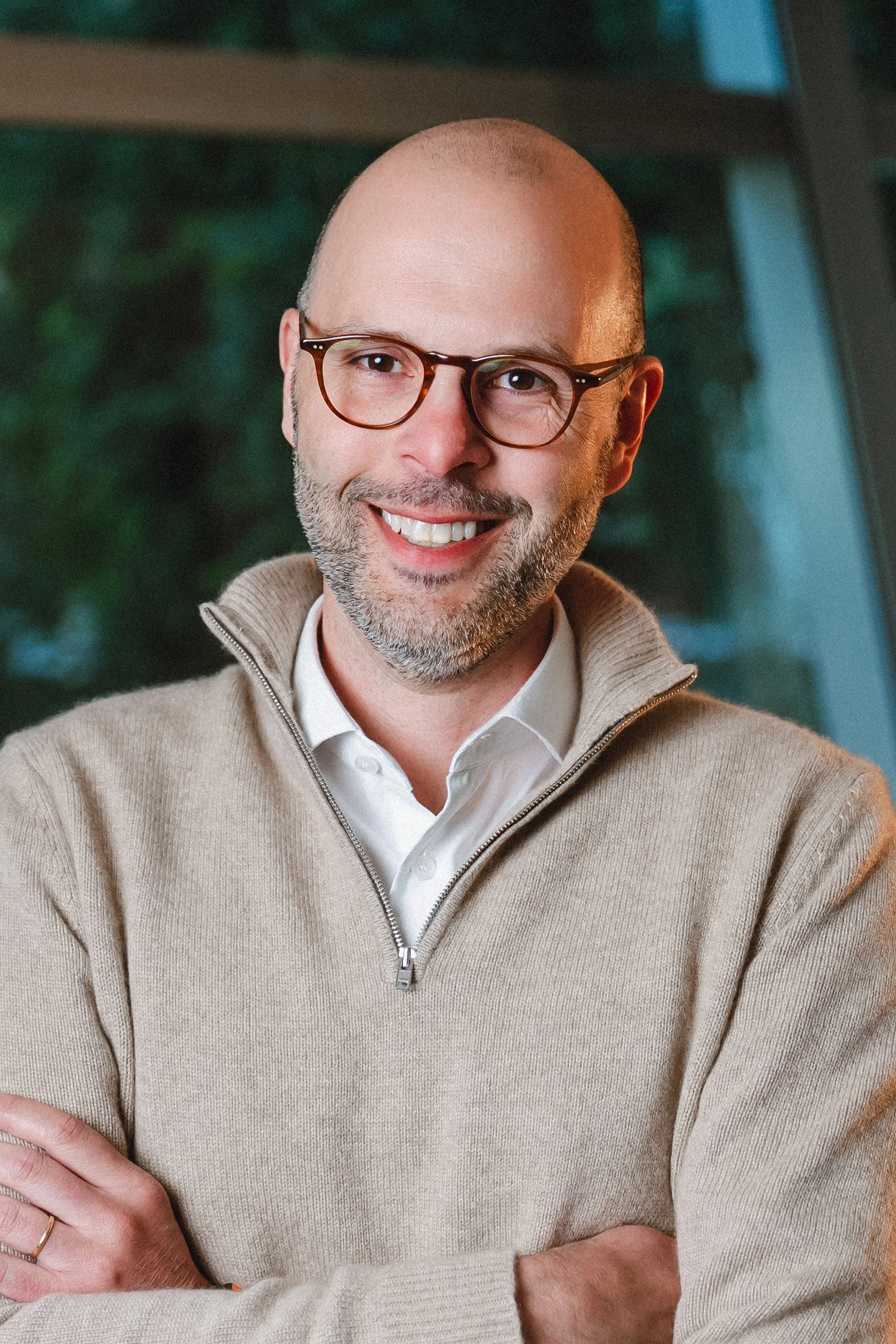 A smiling man with glasses and a beard, wearing a beige zip-up sweater and white collared shirt, standing indoors with blurred green foliage in the background.