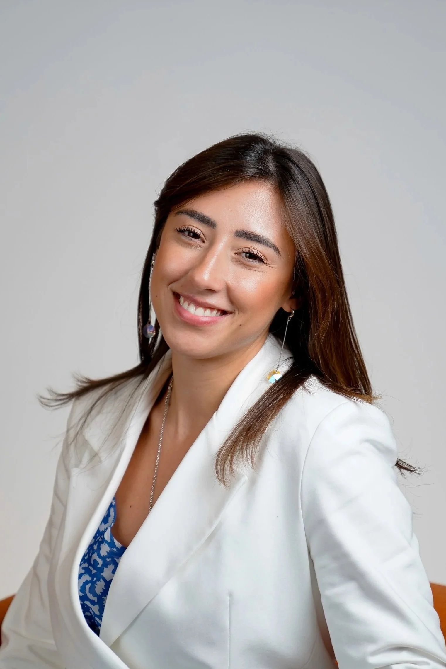 A woman with long brown hair, smiling, wearing a white blazer over a blue patterned top, dangling earrings, and a necklace against a neutral background.