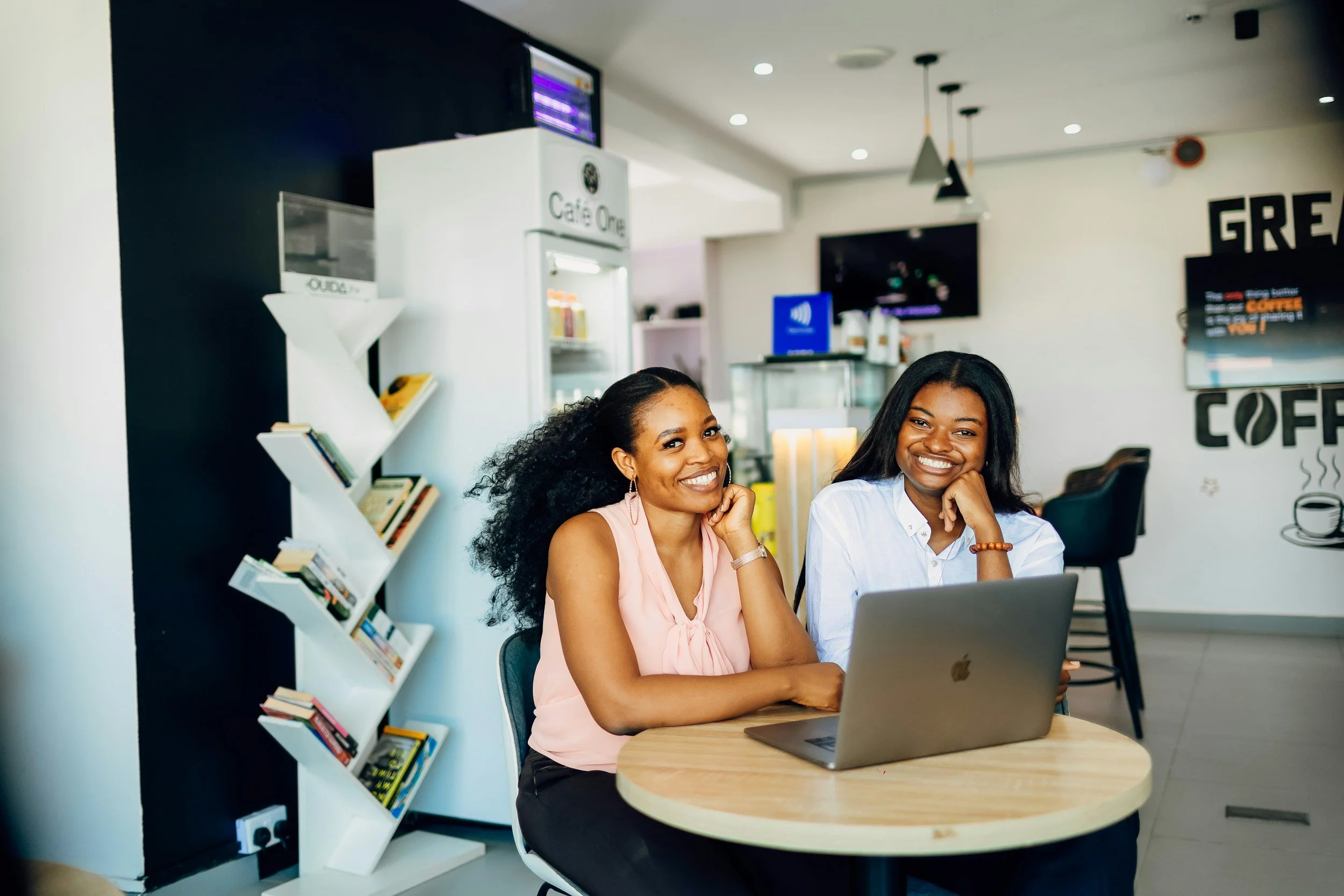 Two women if colour sitting together at a table in front of a laptop computer laughing