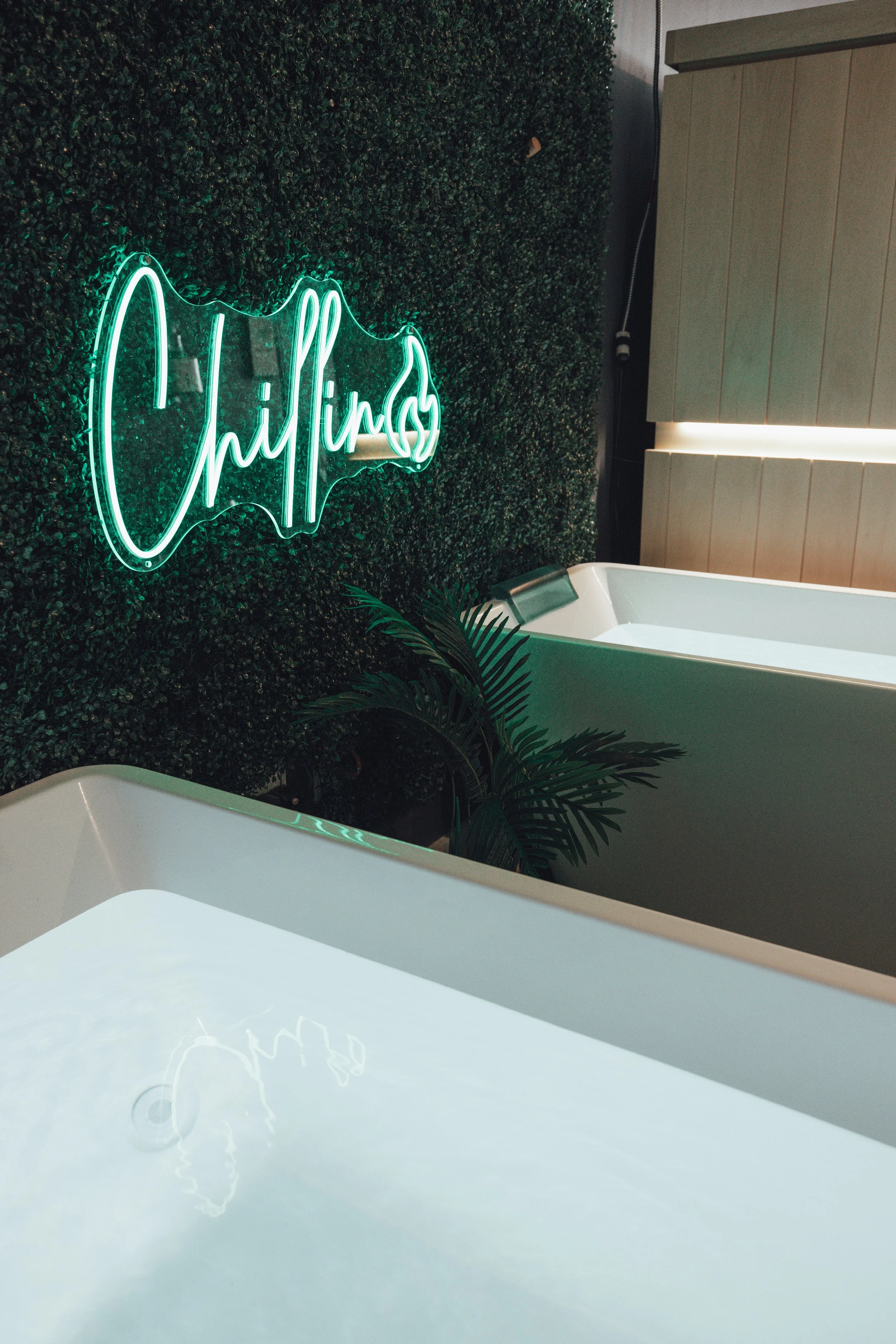 Interior of recovery facility with cold plunge tub, a green artificial hedge wall, a neon sign that says "Chillin'," and some green plants.