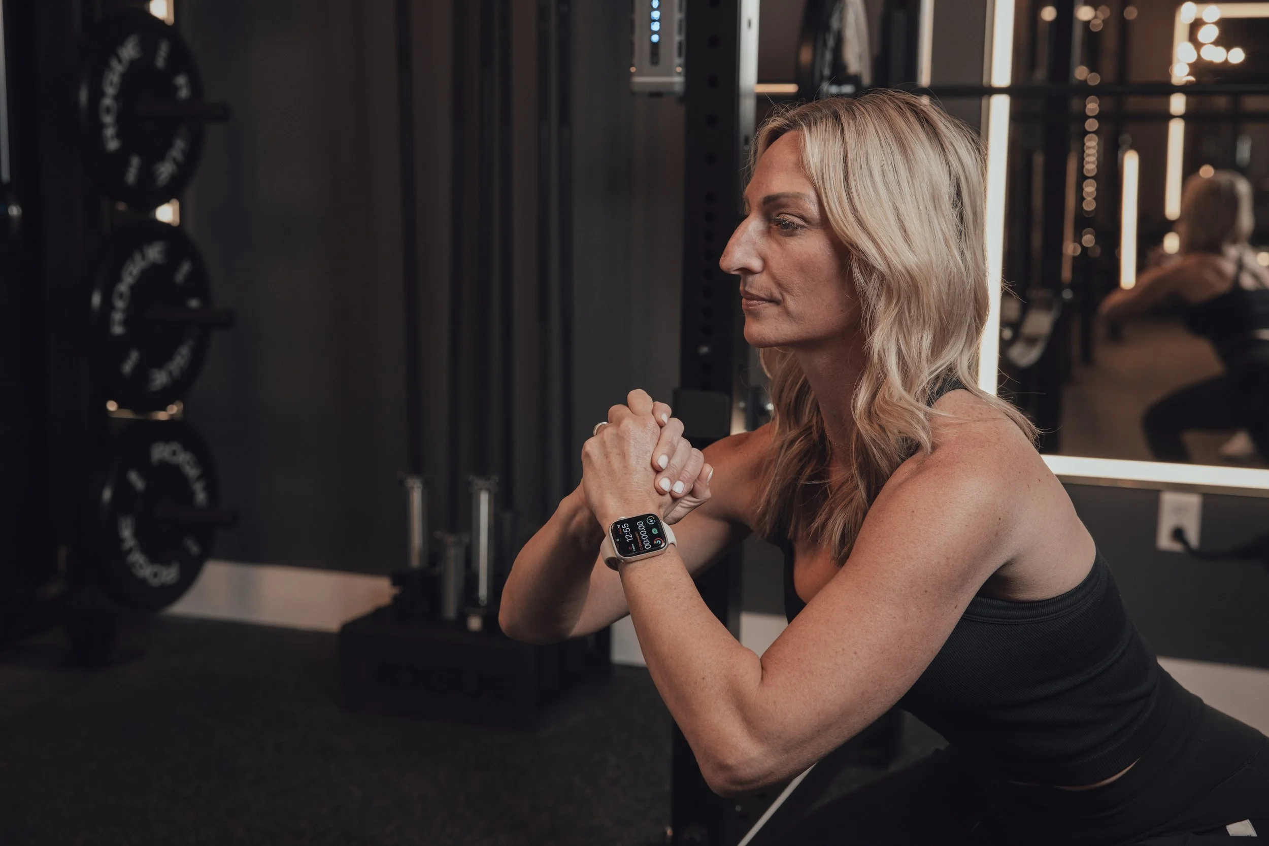 A woman with blonde hair wearing a black athletic top is doing a seated exercise in a gym, looking serenely ahead, with her hands clasped in front of her.
