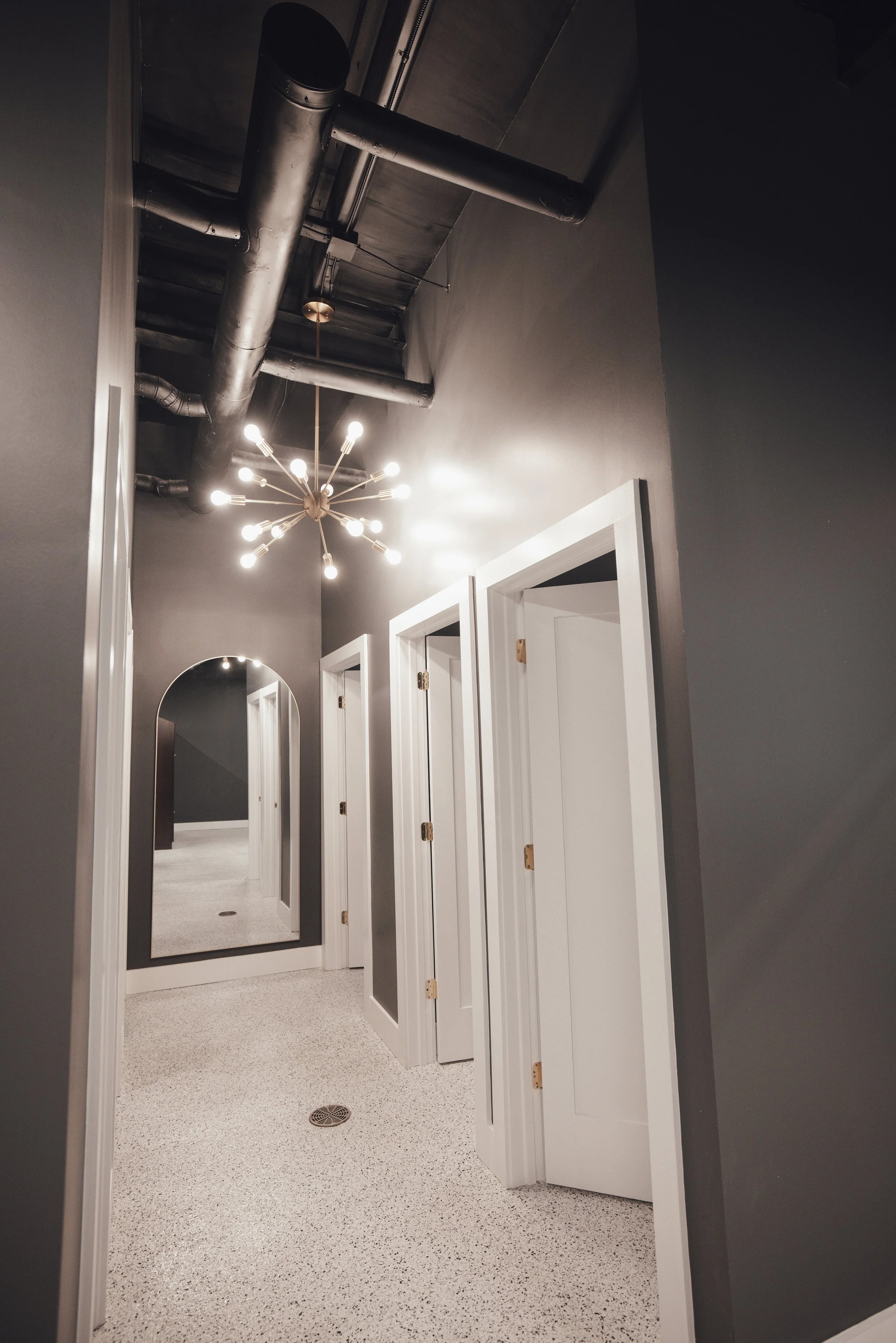 Interior hallway with dark gray walls, white door frames, a large arched mirror, and a modern chandelier with multiple exposed bulbs hanging from the ceiling. Exposed black pipes are visible on the ceiling.