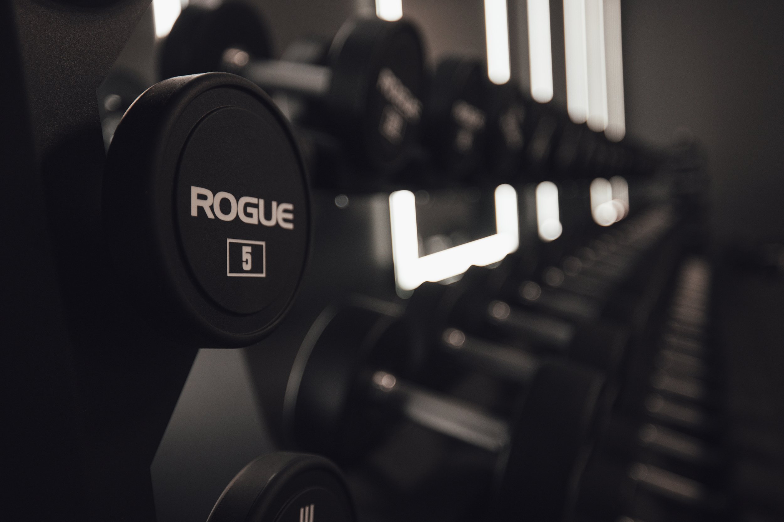 Close-up of black Rogue 5 kettlebell weight on a rack in a gym, with blurred exercise equipment in the background.