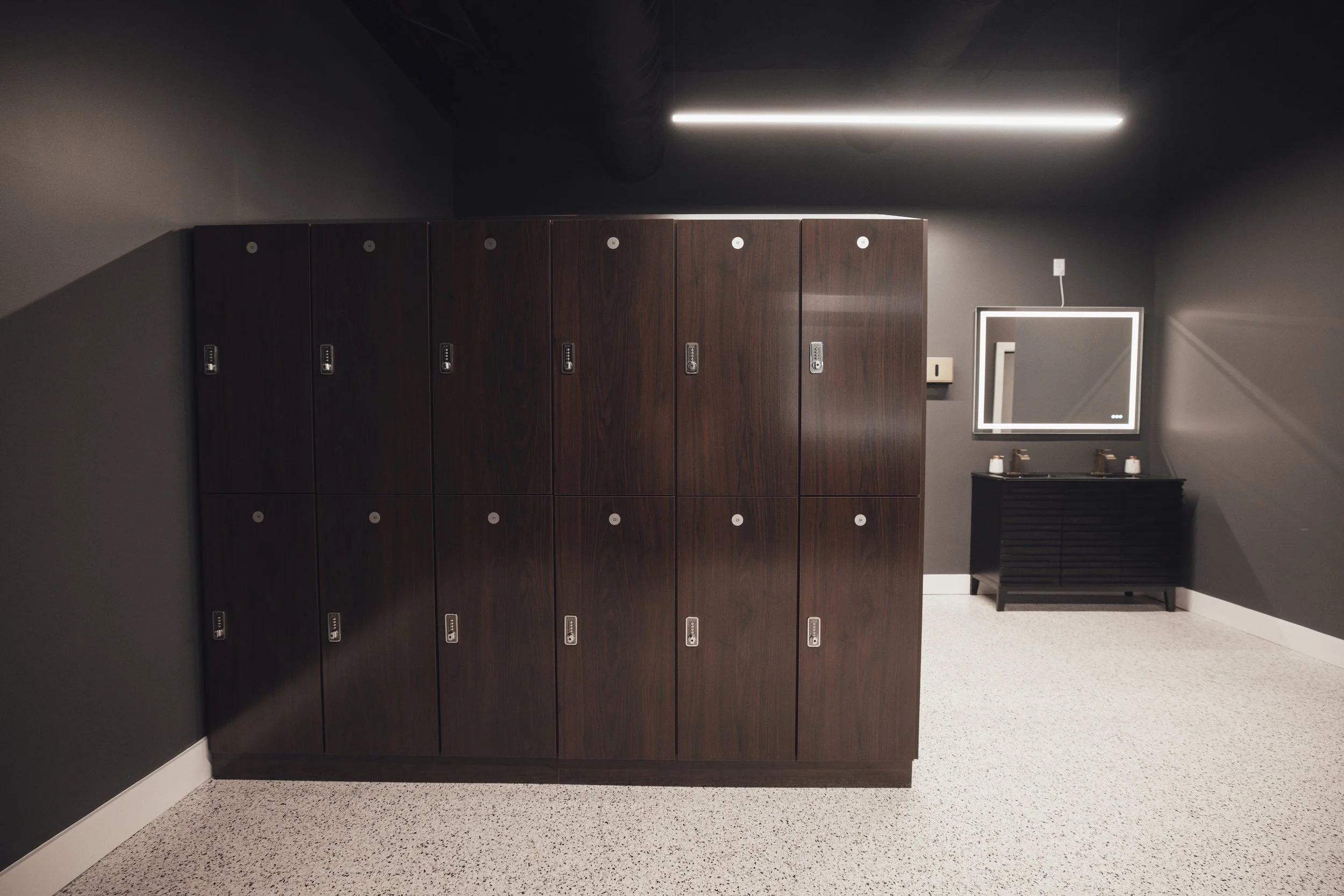 Dark brown lockers next to a black bathroom vanity with a mirror and two sinks in a room with black walls and beige speckled flooring.