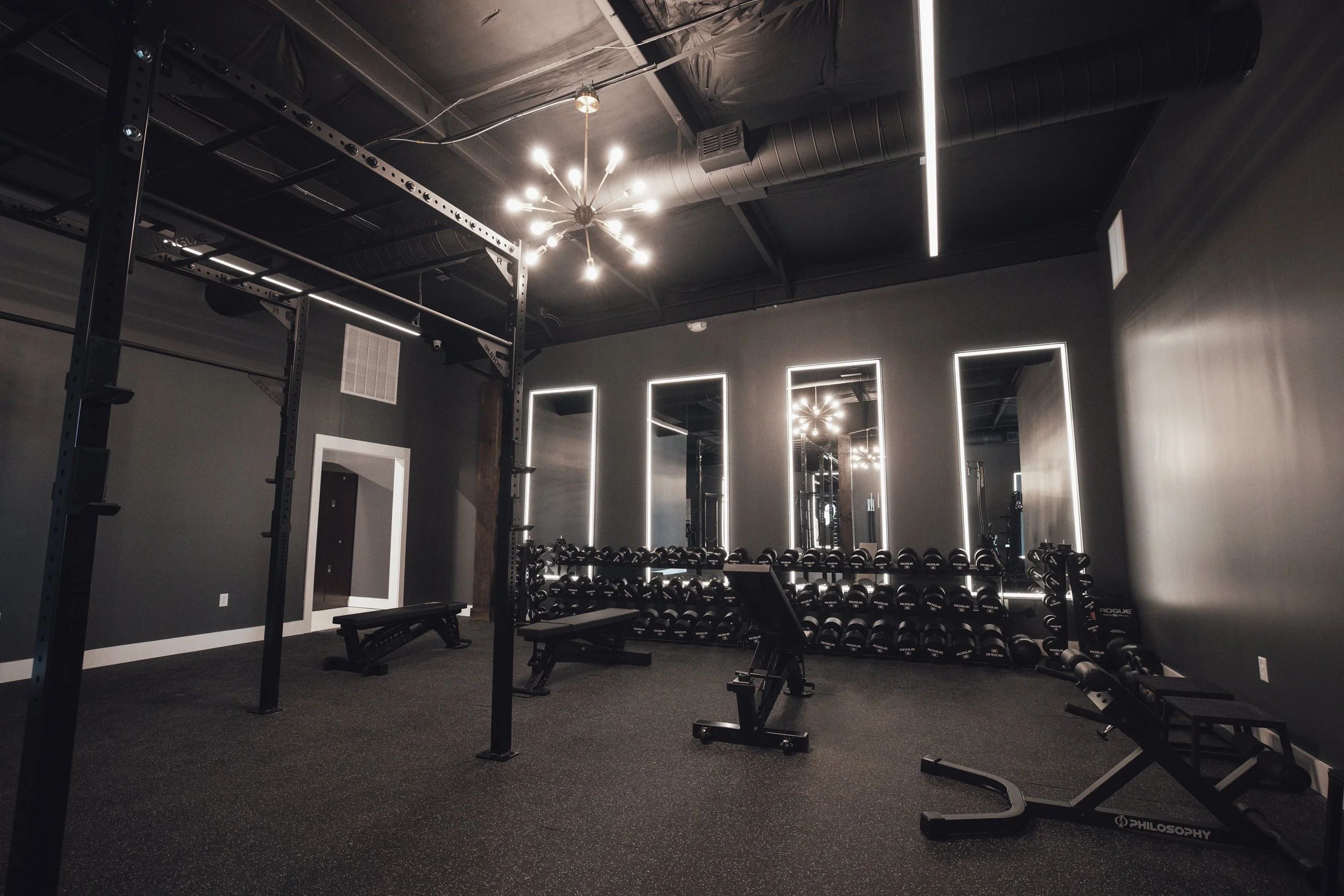 Empty modern gym with black walls, mirrors, and various workout equipment including dumbbells, benches, and a squat rack.