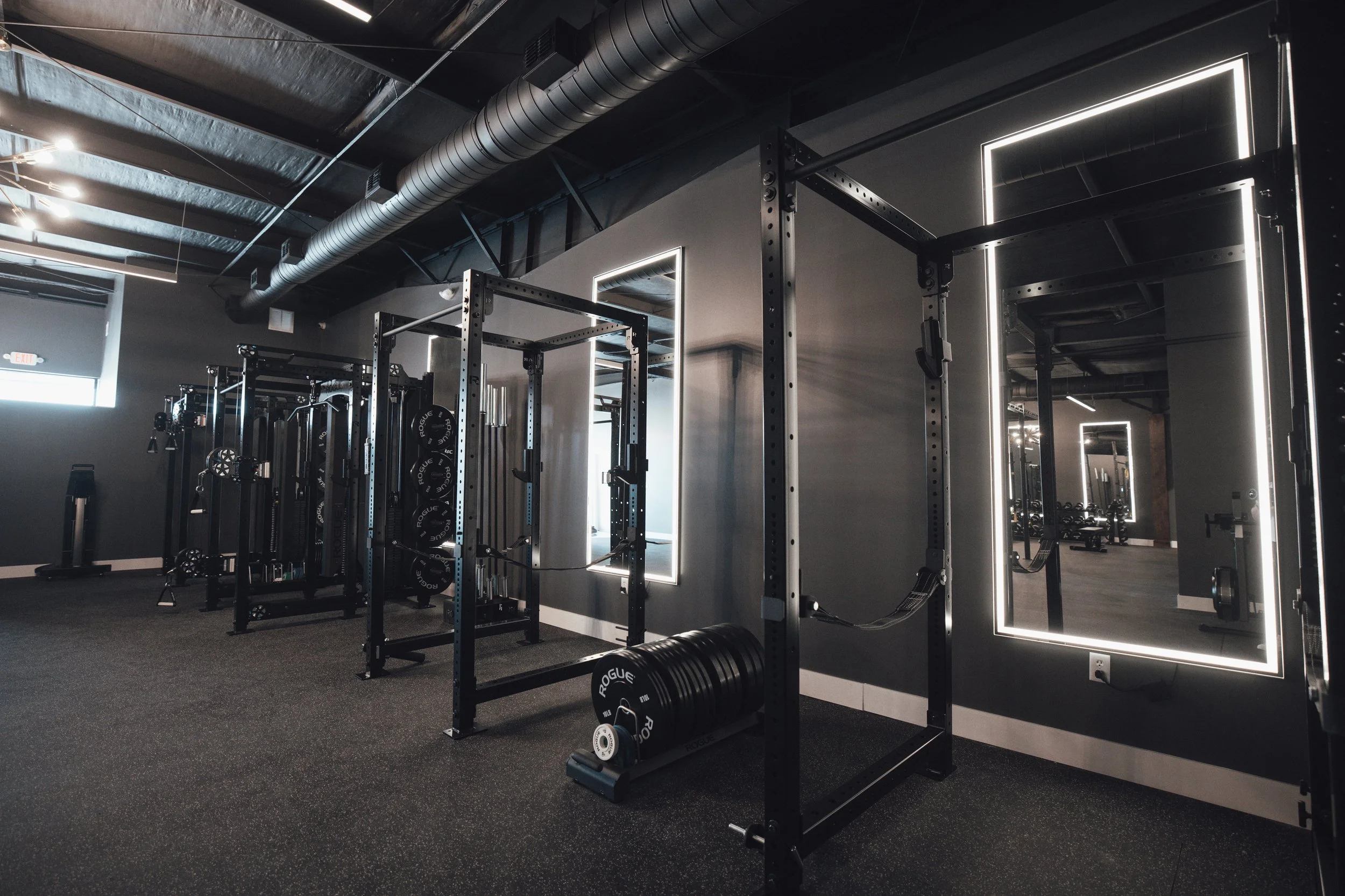 Inside a modern, minimalist gym with black and gray equipment, including a power rack, weight plates, and mirrors with LED lighting, on a dark rubberized floor.