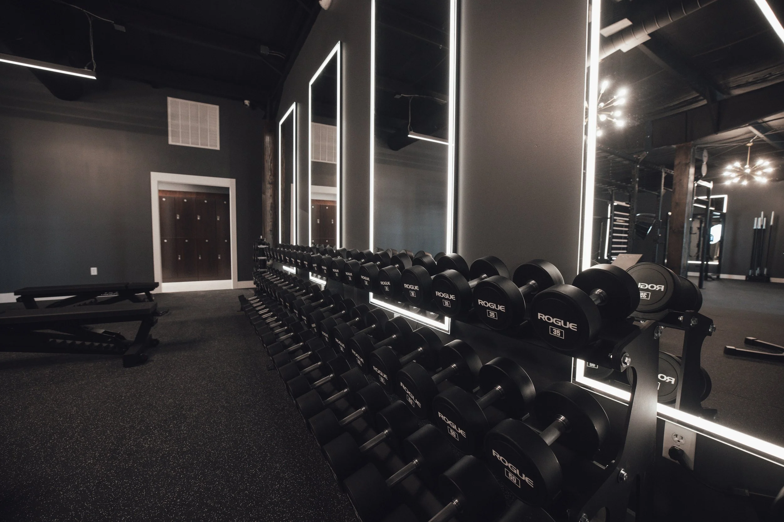 A modern gym with black dumbbells on racks, illuminated mirror panels, and workout benches, with a dark color scheme and ambient lighting.