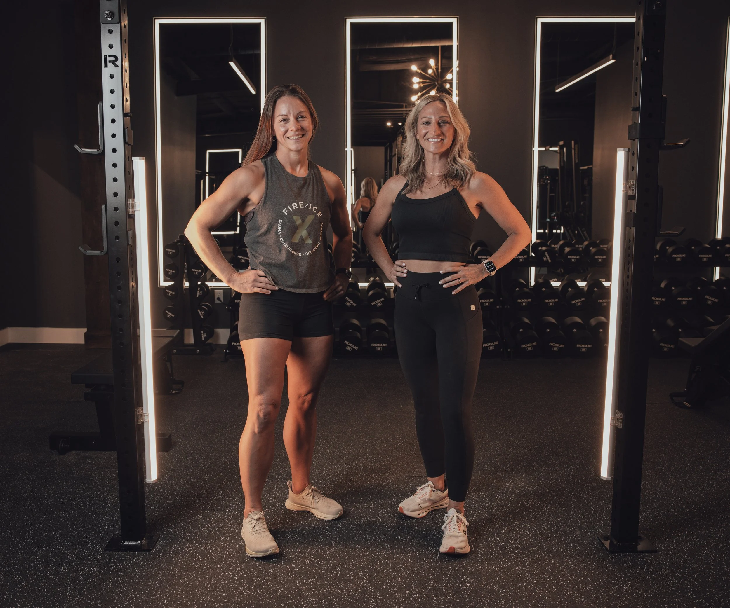 Two women in workout attire standing confidently into the camera inside a modern gym with black walls and illuminated mirrors, fitness equipment in the background.