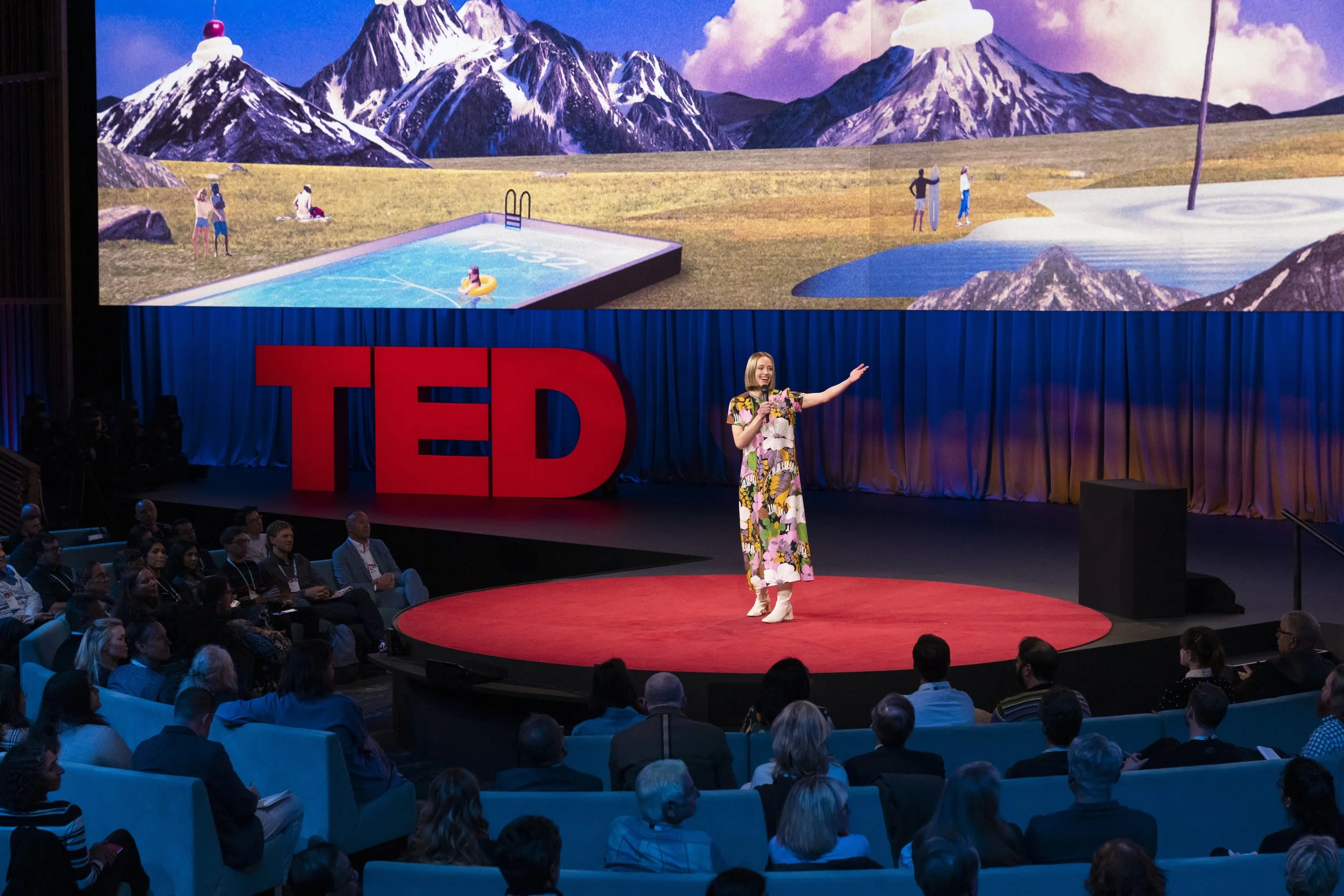 Woman with blonde bob standing on TED stage in floral dress and white boots, gesturing
