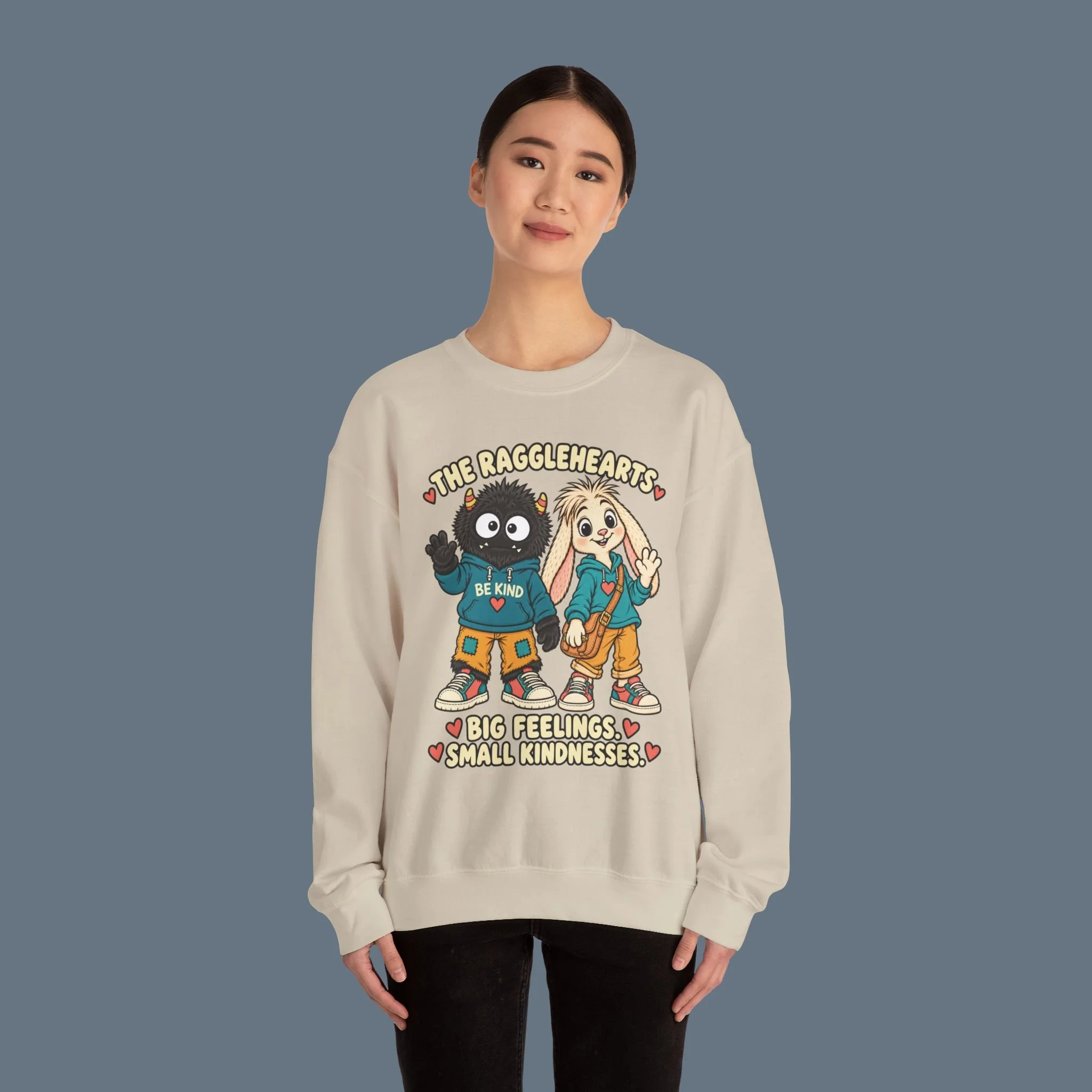 Ragglehearts Sand sweatshirt on a standing model against slate, showing the complete enlarged BIG FEELINGS SMALL KINDNESSES artwork.