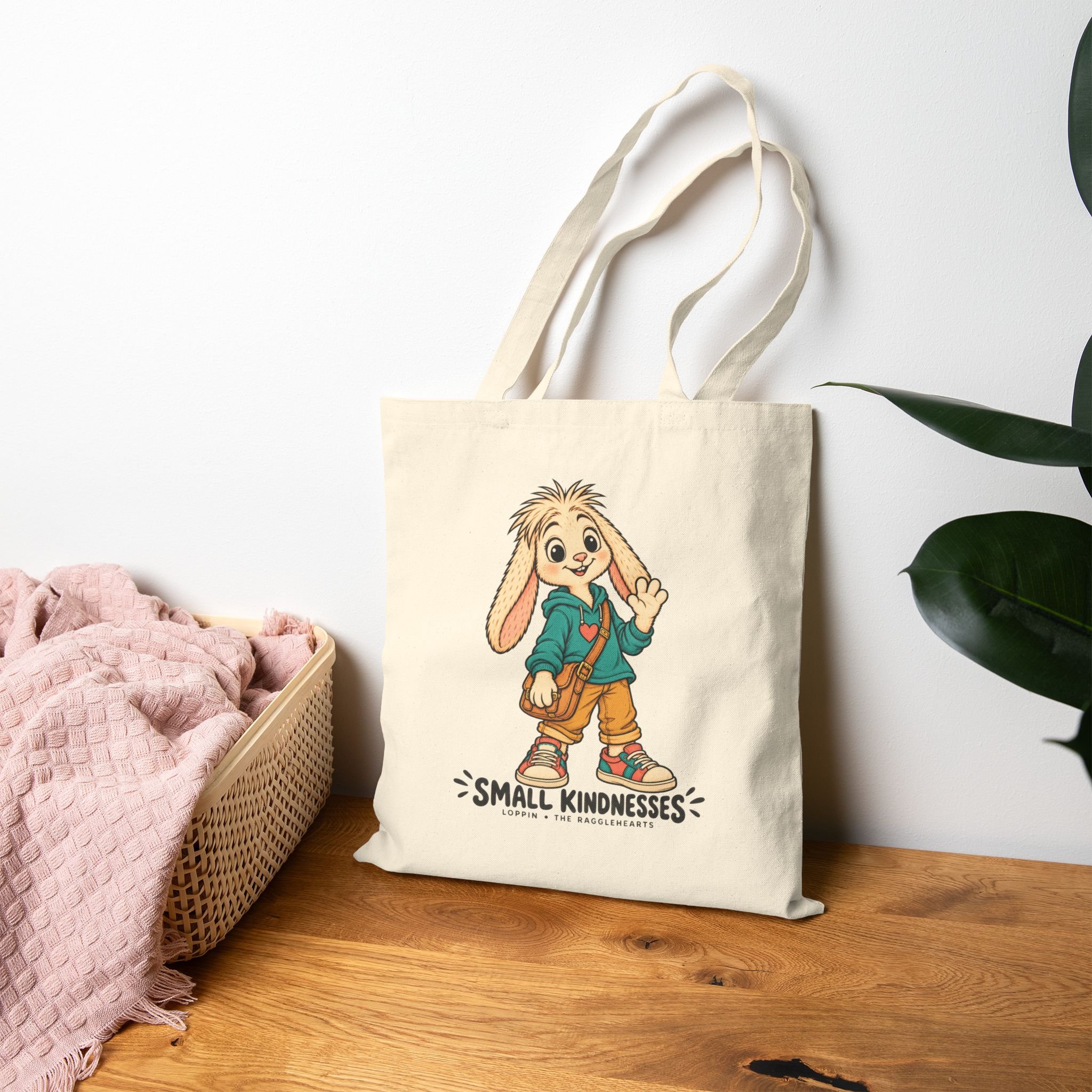 Loppin canvas tote on a wood table, with complete enlarged Small Kindnesses artwork, cream ears, teal hoodie, ochre trousers and tan satchel. Plant and blanket are props.